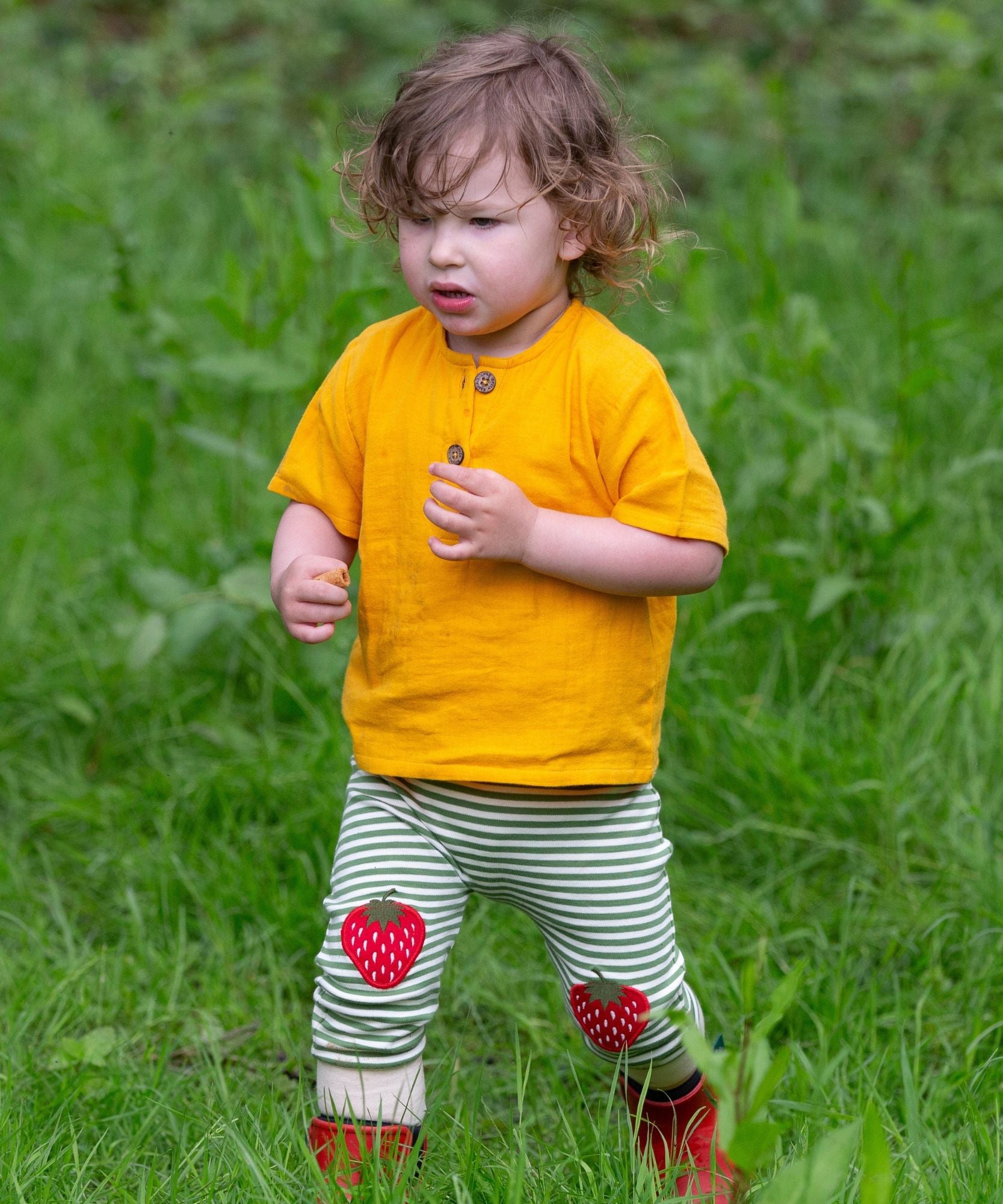 A child wearing the Little Green Radicals Strawberry Knee Patch Green and Cream Striped Organic Cotton Joggers with a bright yellow gold coloured short sleeved t-shirt.