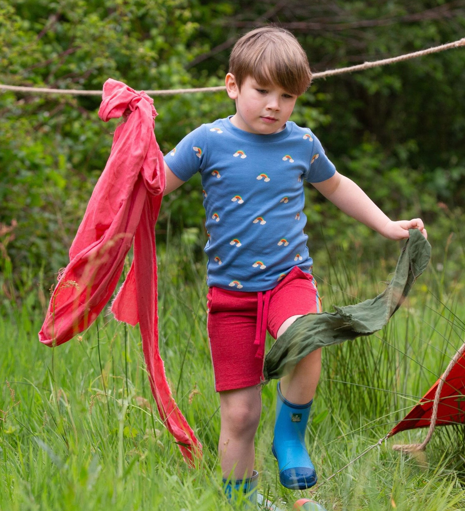 A child wearing the Little Green Radicals Red Comfy Jogger Shorts with a blue rainbow short sleeved t-shirt. The child is running through long green grass.