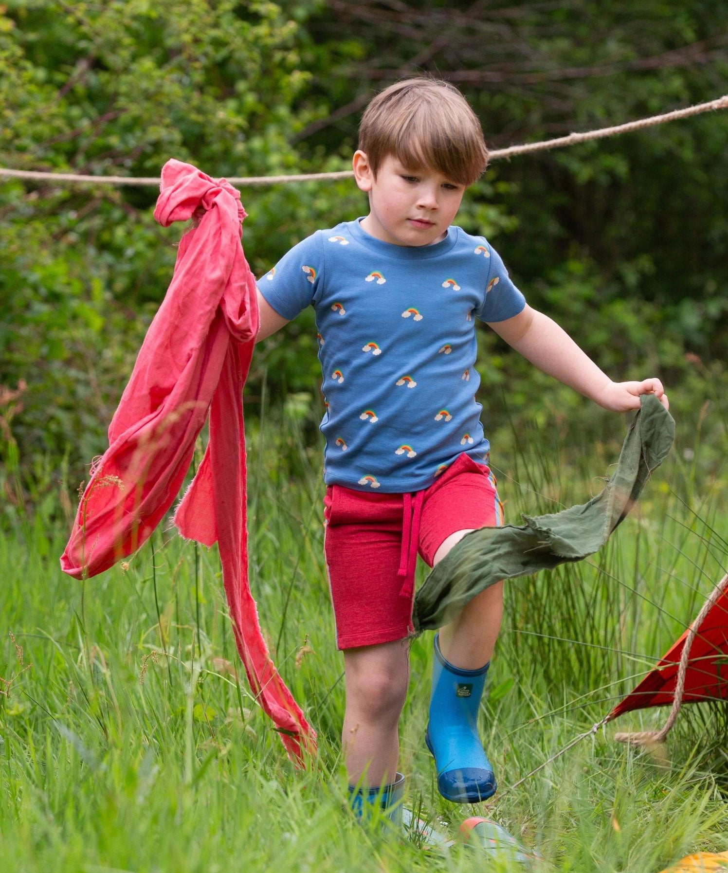 A child wearing the Little Green Radicals Red Comfy Jogger Shorts with a blue rainbow short sleeved t-shirt. The child is running through long green grass.