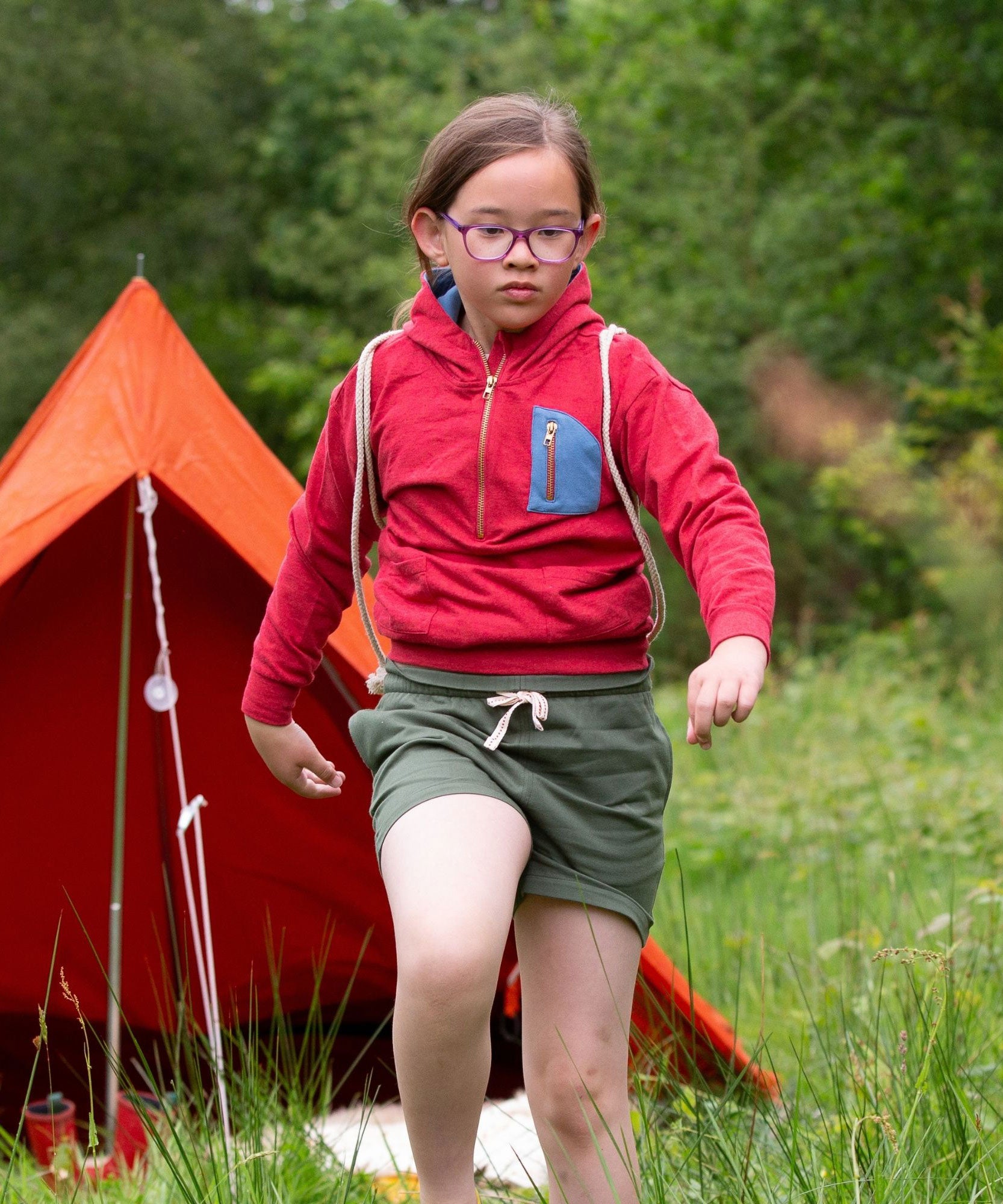 A child wearing the Little Green Radicals Child's Red Organic Quarter Zip Hoodie with green shorts. An orange tenet can be seen in the back ground.