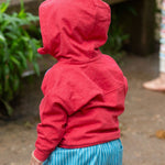 A child wearing the Little Green Radicals Child's Red Organic Quarter Zip Hoodie with blue striped shorts. The child is facing away from the camera with the hood of the top up 