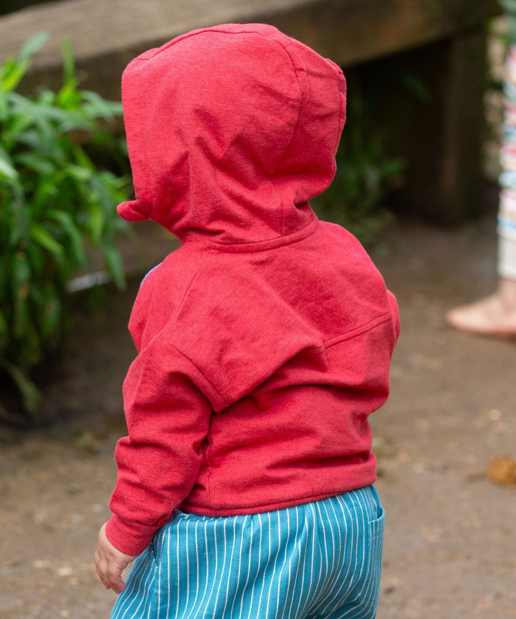 A child wearing the Little Green Radicals Child's Red Organic Quarter Zip Hoodie with blue striped shorts. The child is facing away from the camera with the hood of the top up 