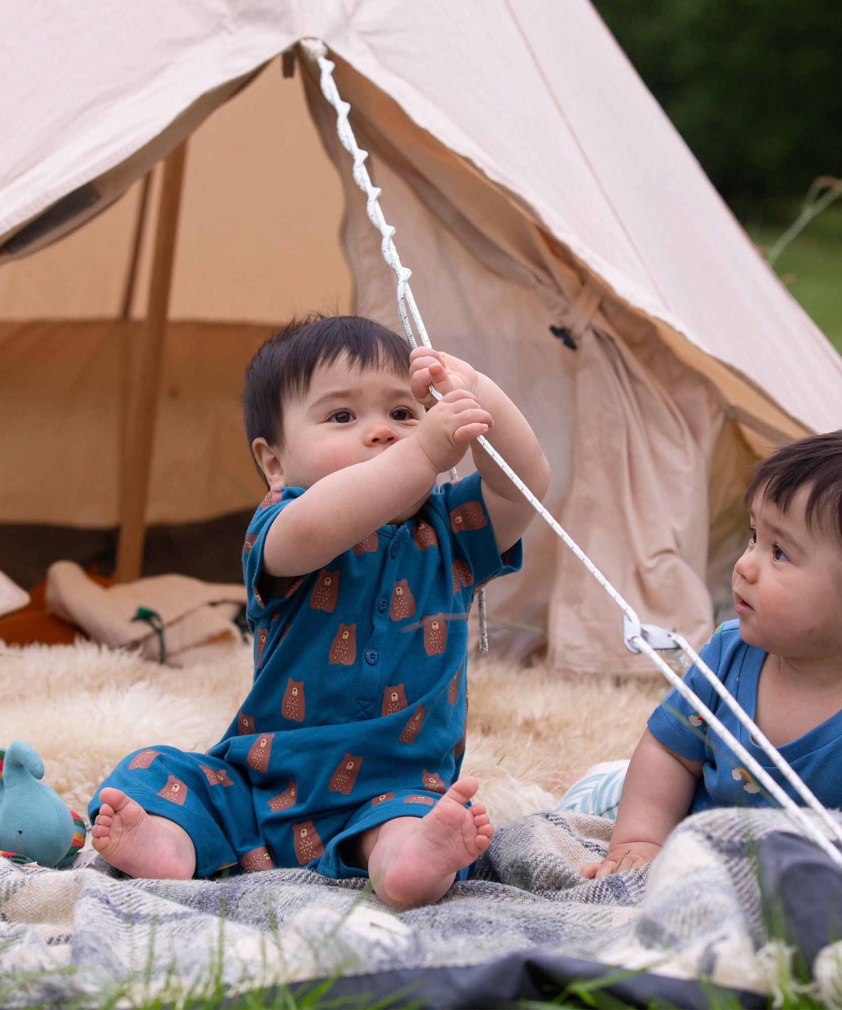 A child sitting in front of a bell tent wearing the Little Green Radicals Teddy Bears Organic Cotton Shortie Romper. 