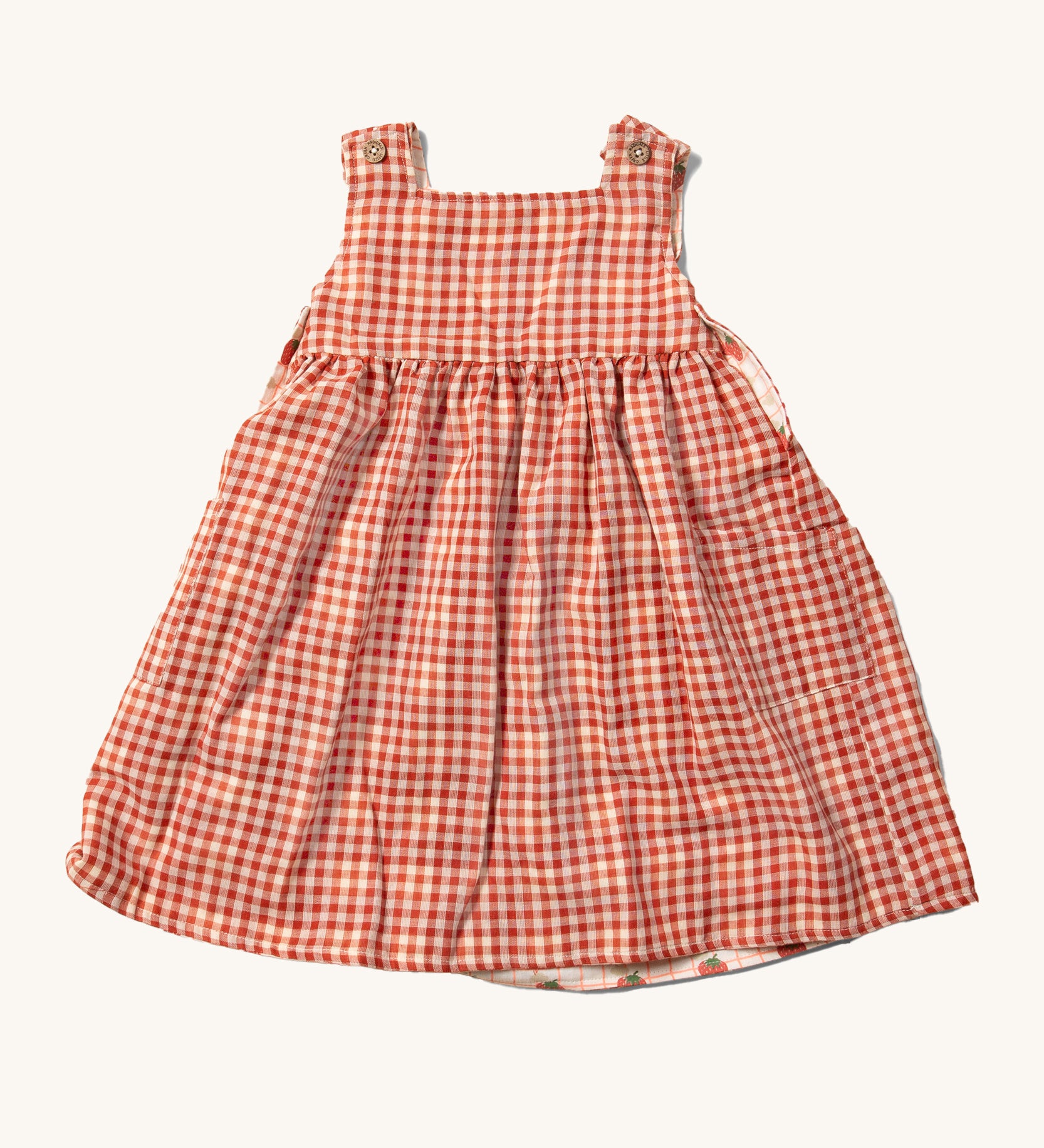 Little Green Radicals Child's Organic Cotton Strawberry Days Reversible Pinny Dress shown with the red gingham side on the outside.