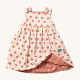 Little Green Radicals Child's Organic Cotton Strawberry Days Reversible Pinny Dress shown with the strawberry print on the outside. The corner of the dress is show lifted with the red gingham print on the other side showing.