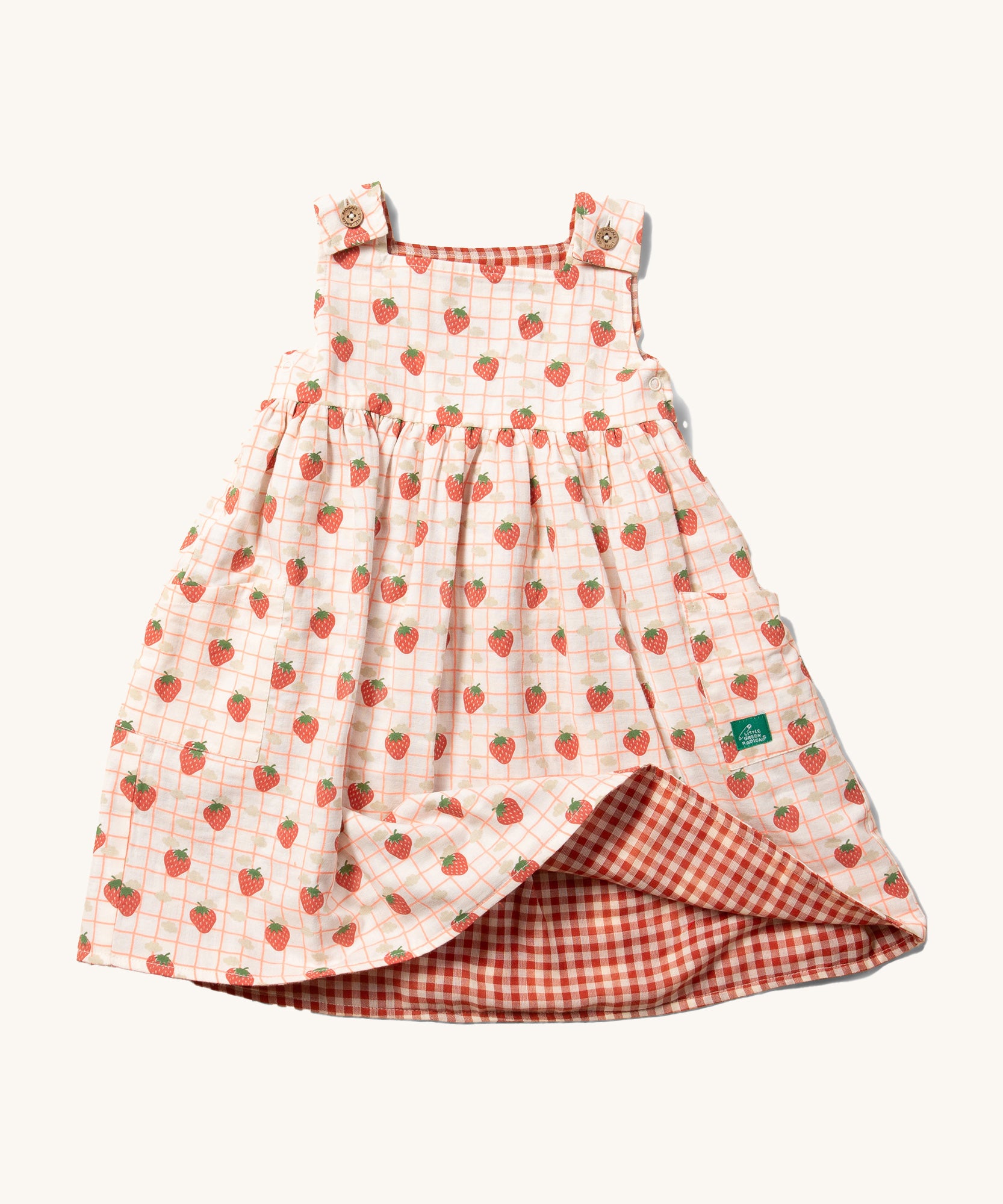 Little Green Radicals Child's Organic Cotton Strawberry Days Reversible Pinny Dress shown with the strawberry print on the outside. The corner of the dress is show lifted with the red gingham print on the other side showing.