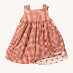 Little Green Radicals Child's Organic Cotton Strawberry Days Reversible Pinny Dress shown with the red gingham on the outside. The corner of the dress is show lifted with the strawberry print on the other side showing.