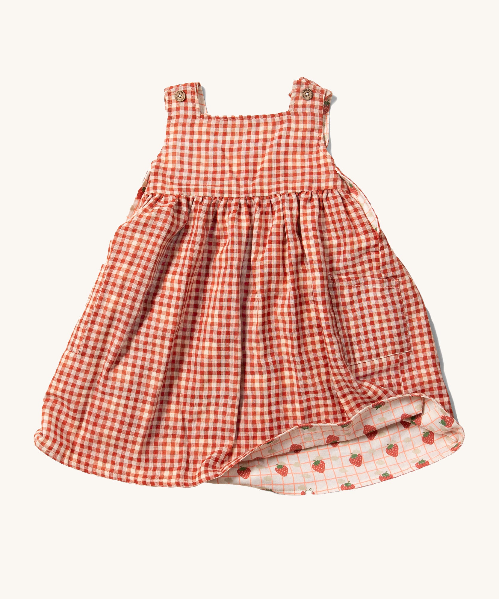 Little Green Radicals Child's Organic Cotton Strawberry Days Reversible Pinny Dress shown with the red gingham on the outside. The corner of the dress is show lifted with the strawberry print on the other side showing.