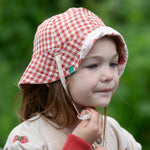 A child wearing the Little Green Radicals Strawberry Days Reversible Kids Sun Hat. The child is wearing it with the red and cream gingham side showing on the outside.