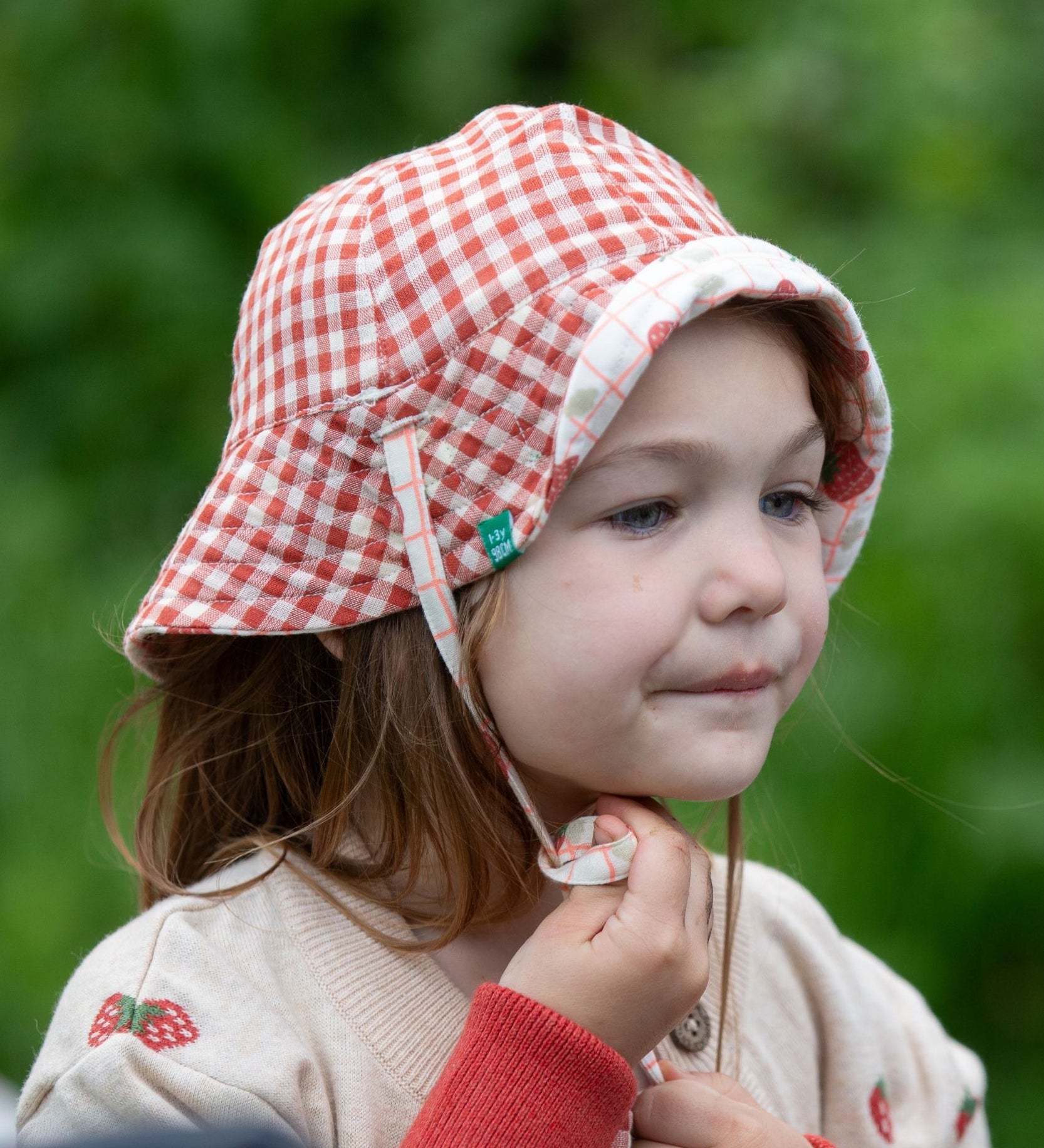 A child wearing the Little Green Radicals Strawberry Days Reversible Kids Sun Hat. The child is wearing it with the red and cream gingham side showing on the outside.