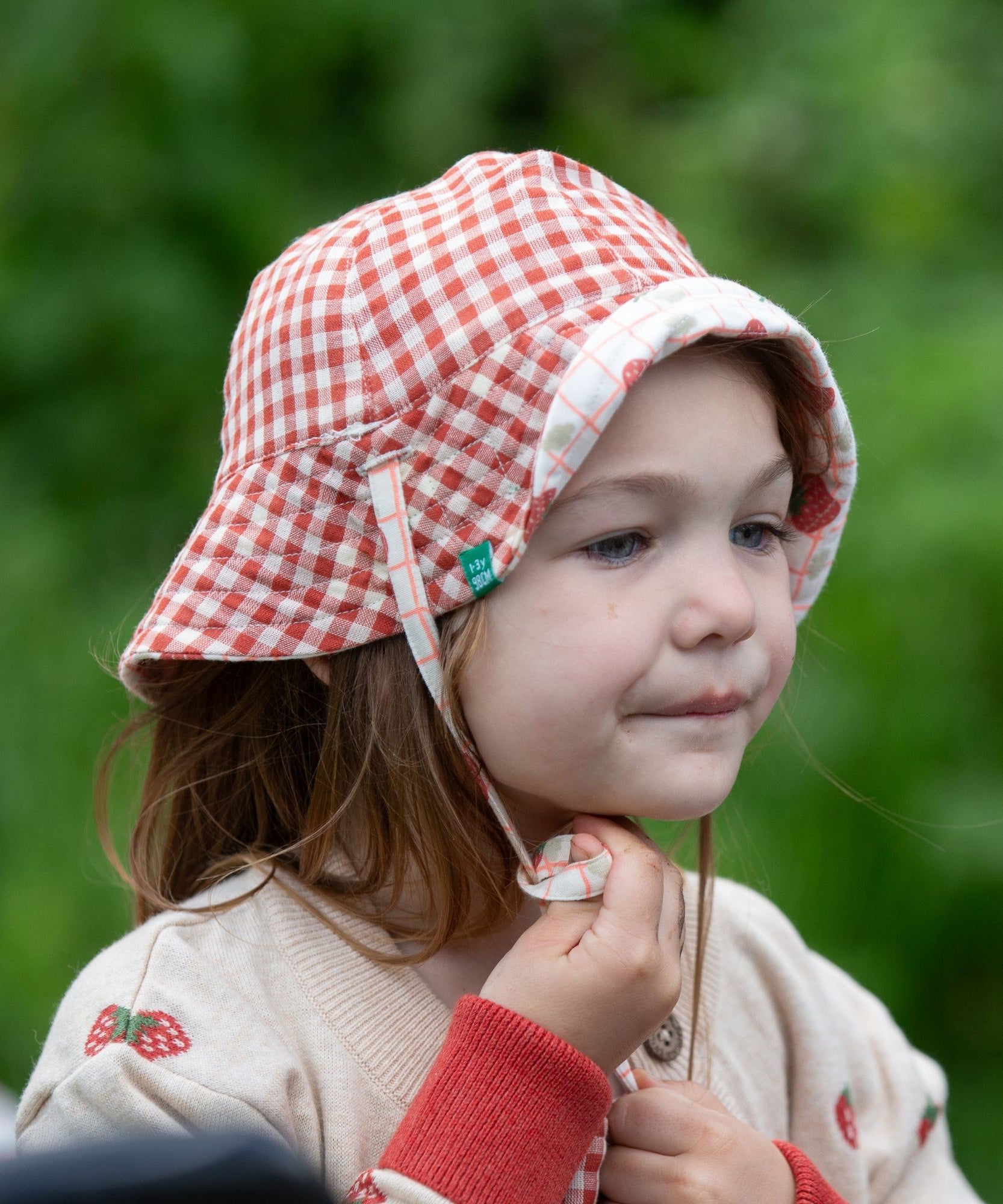 A child wearing the Little Green Radicals Strawberry Days Reversible Kids Sun Hat. The child is wearing it with the red and cream gingham side showing on the outside.