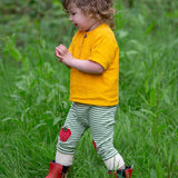 A child wearing the Little Green Radicals Strawberry Knee Patch Green and Cream Striped Organic Cotton Joggers with a bright yellow gold coloured short sleeved t-shirt.