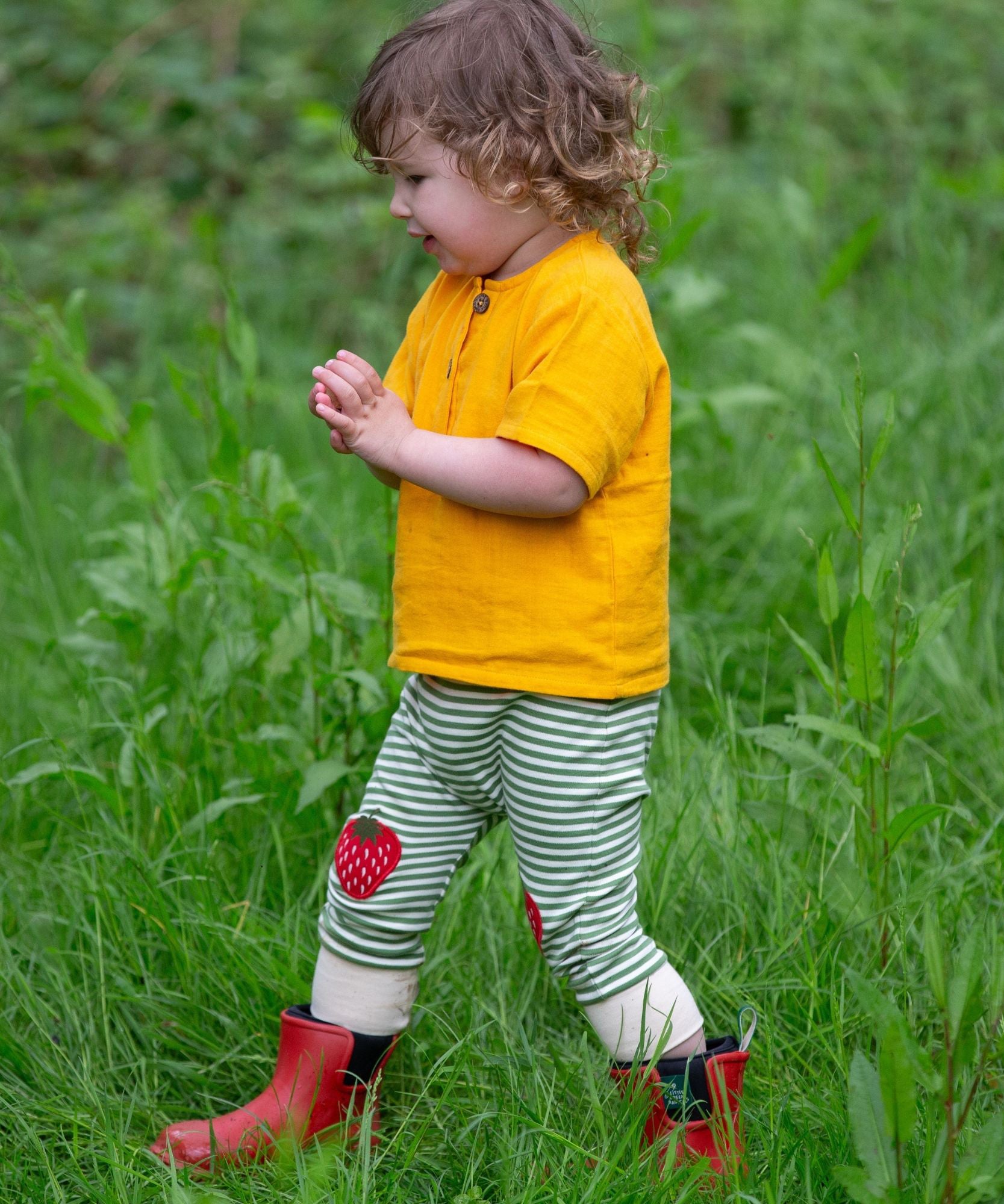 A child wearing the Little Green Radicals Strawberry Knee Patch Green and Cream Striped Organic Cotton Joggers with a bright yellow gold coloured short sleeved t-shirt.