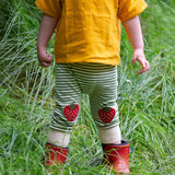 A close up of a child wearing the Little Green Radicals Strawberry Knee Patch Green and Cream Striped Organic Cotton Joggers with a bright yellow gold coloured short sleeved t-shirt.