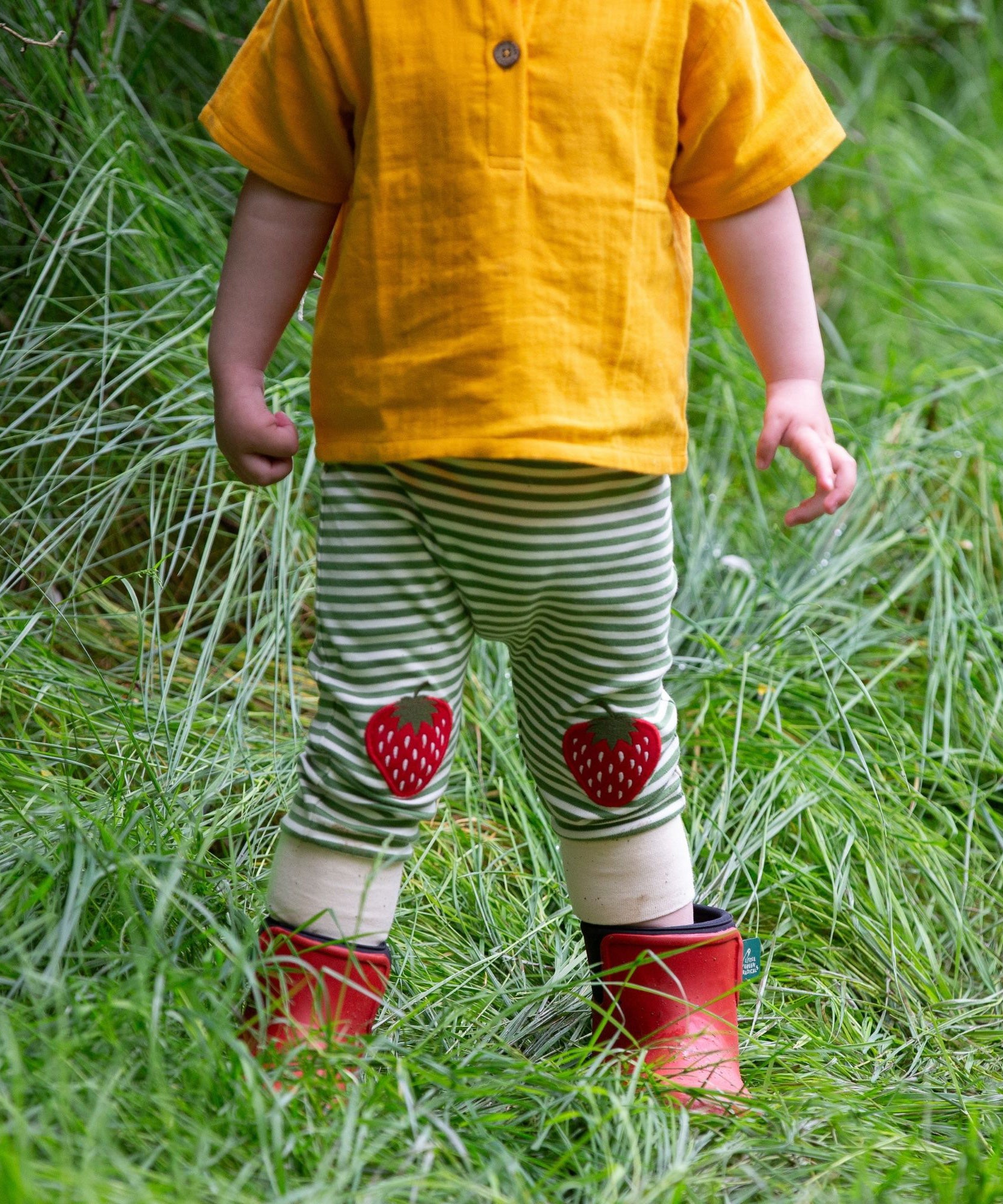 A close up of a child wearing the Little Green Radicals Strawberry Knee Patch Green and Cream Striped Organic Cotton Joggers with a bright yellow gold coloured short sleeved t-shirt.