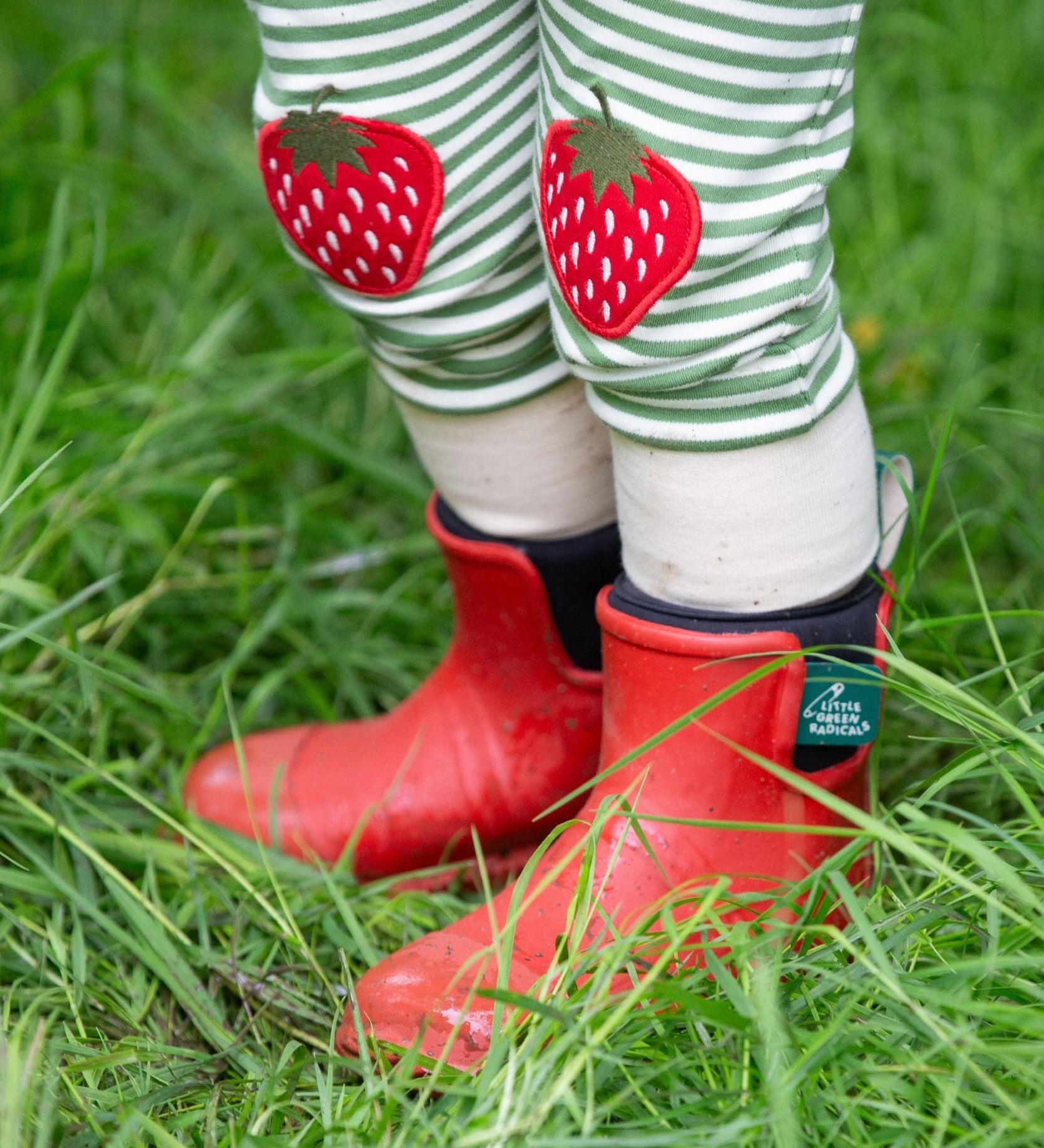 A close up of a child wearing the Little Green Radicals Children's Strawberry Knee Patch Green and Cream Striped Organic Cotton Joggers with red wellington boots