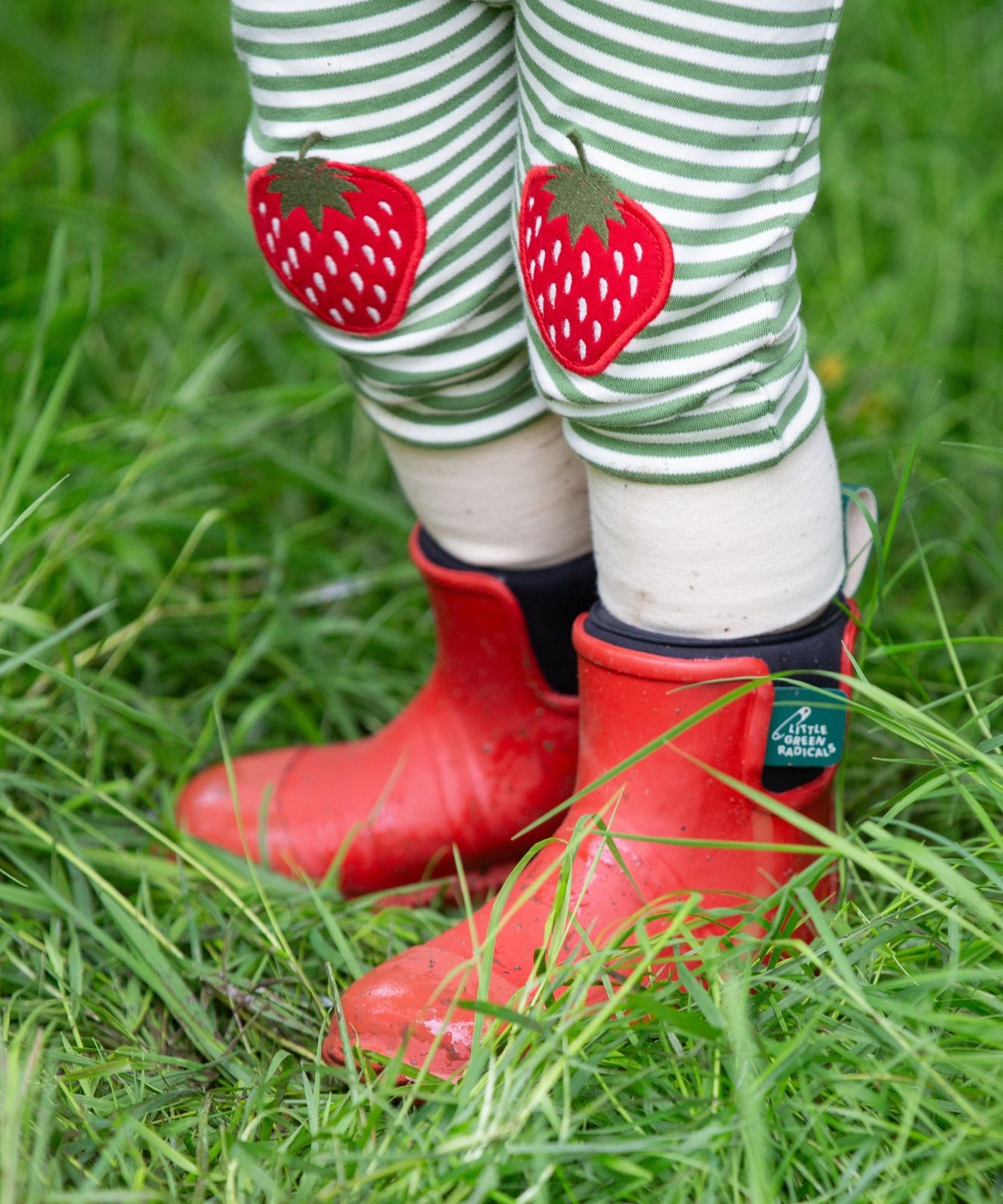 A close up of a child wearing the Little Green Radicals Children's Strawberry Knee Patch Green and Cream Striped Organic Cotton Joggers with red wellington boots