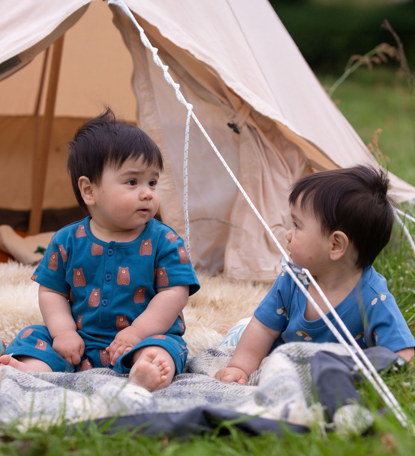 A child sitting in front of a bell tent wearing the Little Green Radicals Teddy Bears Organic Cotton Shortie Romper. 