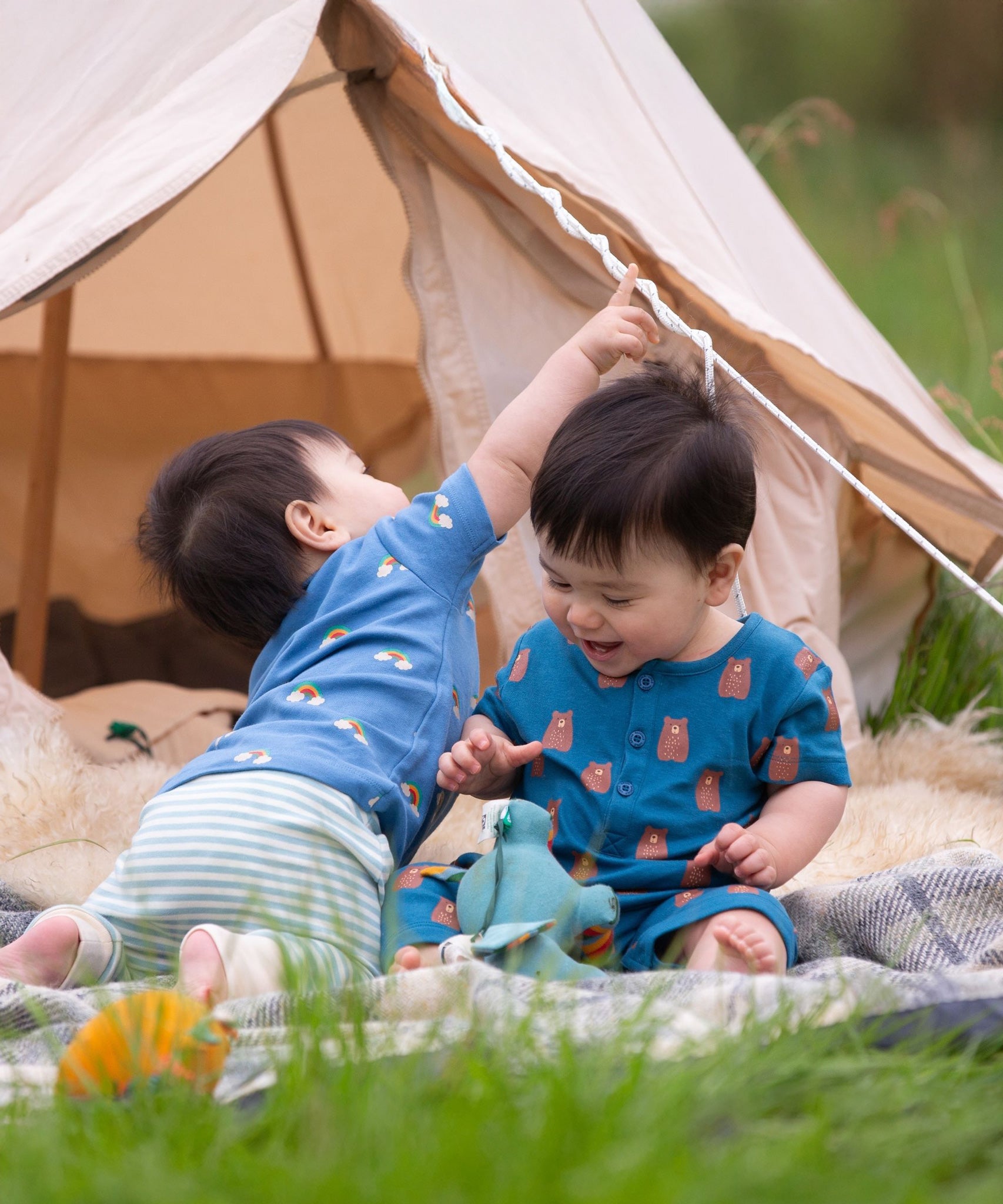 Two young children sitting in front of a bell tent one is wearing the Little Green Radicals Teddy Bears Organic Cotton Shortie Romper the other is wearing the Blue Rainbow playset. 