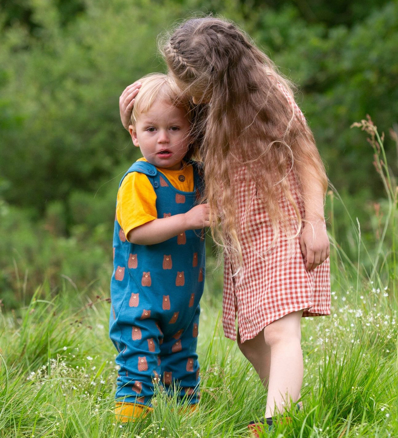 Two children standing in grass,the younger child is wearing the Little Green Radicals Everyday Dungarees with a Teddy Bears print, the older child is wearing a red and white gingham dress.