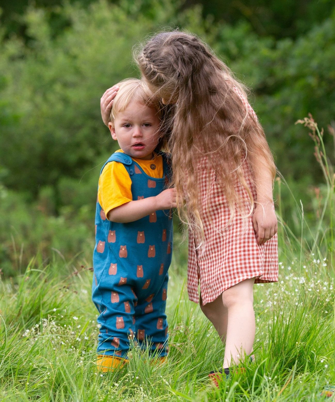 Two children standing in grass,the younger child is wearing the Little Green Radicals Everyday Dungarees with a Teddy Bears print, the older child is wearing a red and white gingham dress.