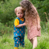 Two children standing in grass,the younger child is wearing the Little Green Radicals Everyday Dungarees with a Teddy Bears print, the older child is wearing a red and white gingham dress.