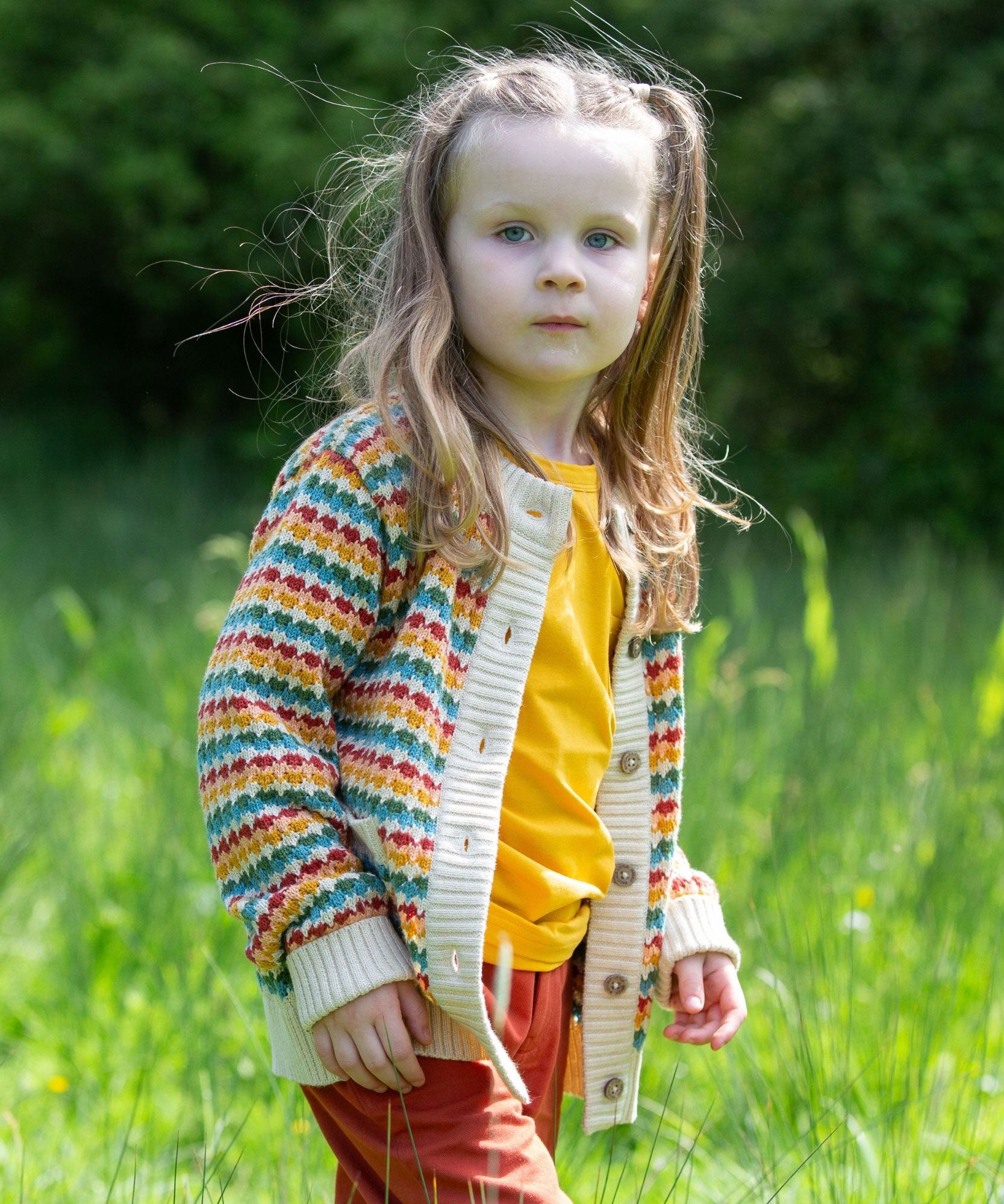 A child wearing the Little Green Radicals From One To Another Rainbow Days Organic Cotton Knitted Cardigan with red shorts.