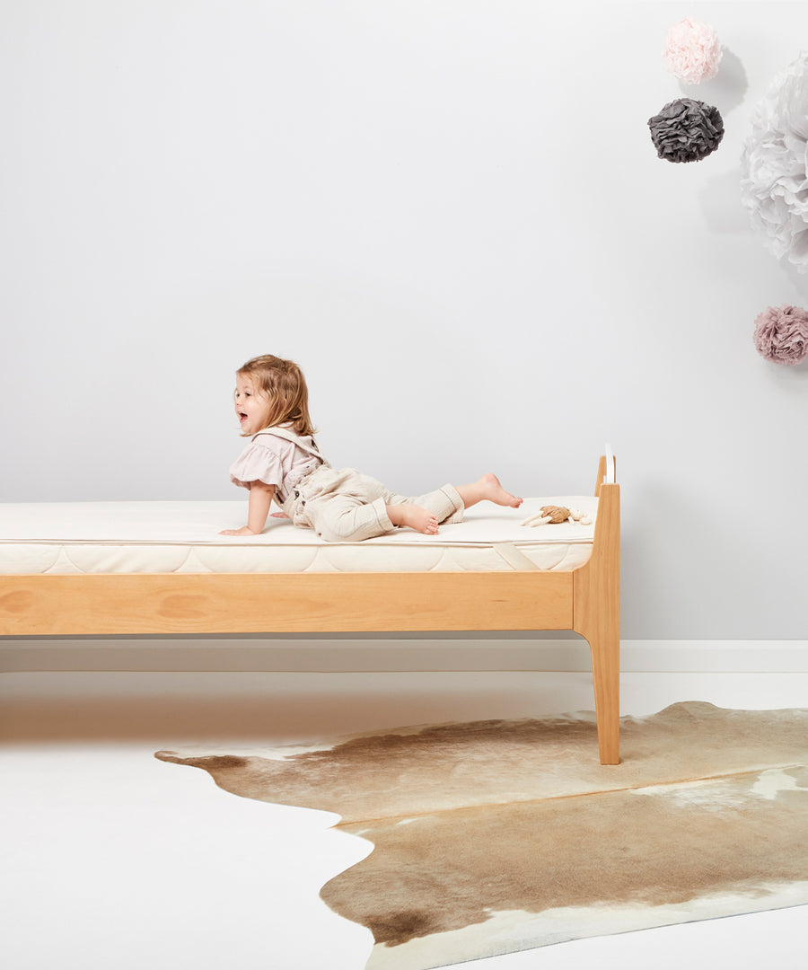 A child is seen lying on a bed which has a Little Green Sheep Organic Junior Mattress Protector placed on the matress