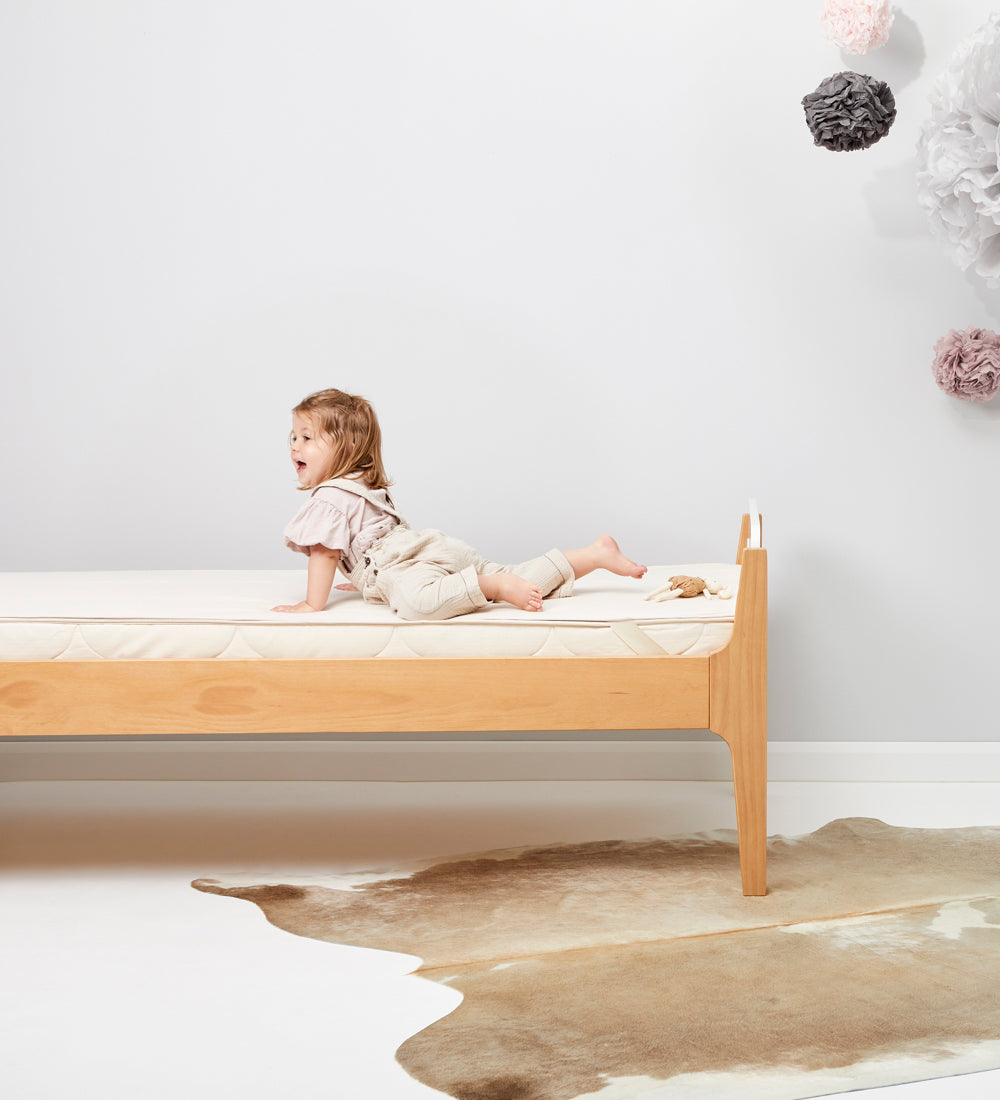 A child is seen lying on a bed which has a Little Green Sheep Organic Junior Mattress Protector placed on the matress