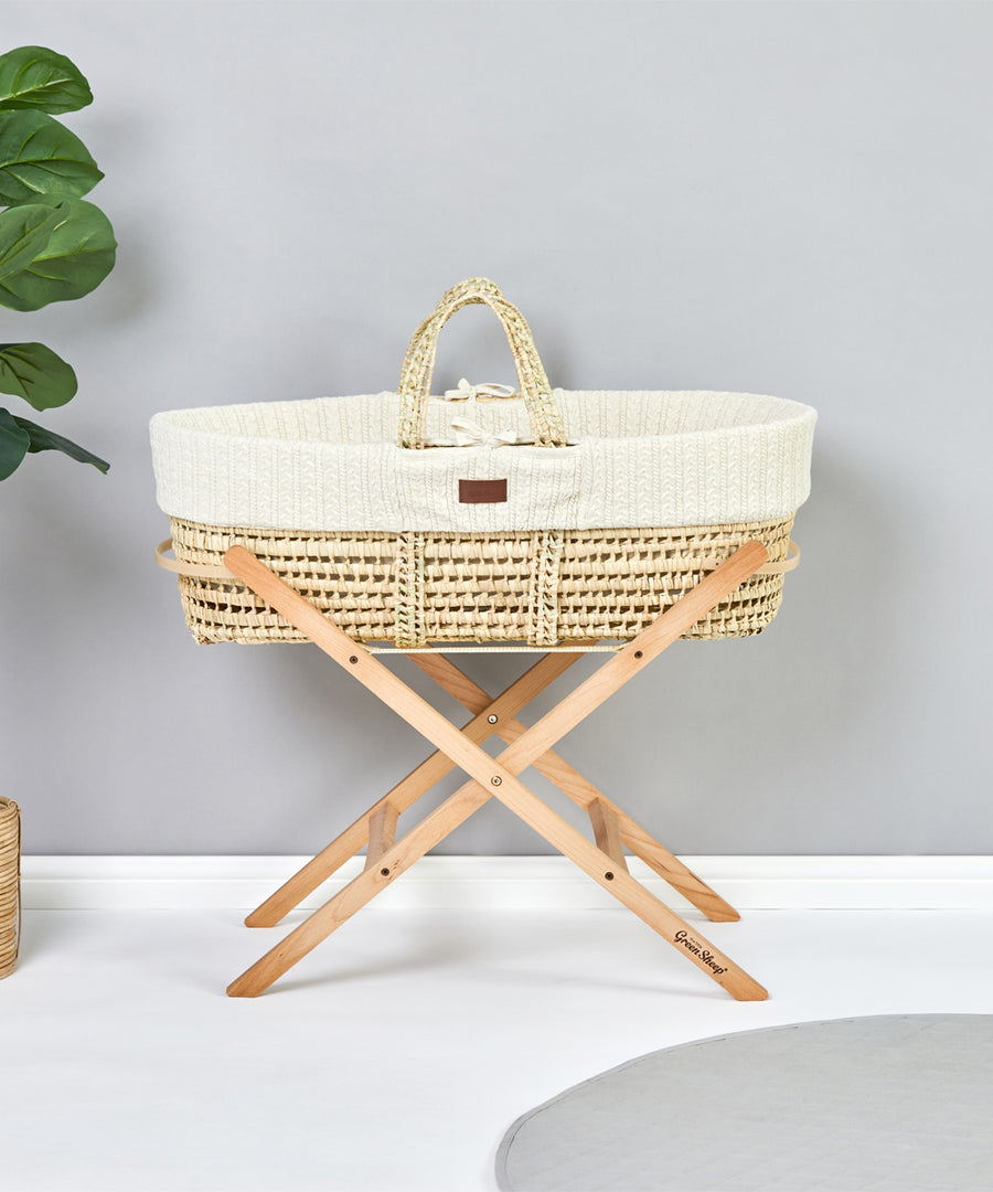 Little Green Sheep Moses Basket Stand pictured in a room with a grey coloured wall. A moses basket with a knitted insert is on the stand