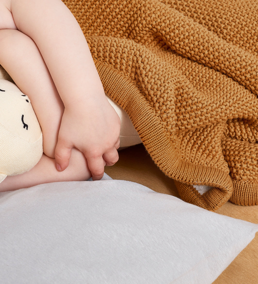 A close up of a child's hands poking out from underneath a honey yellow coloured Little Green Sheep Organic Knitted Fleece Baby Blanket