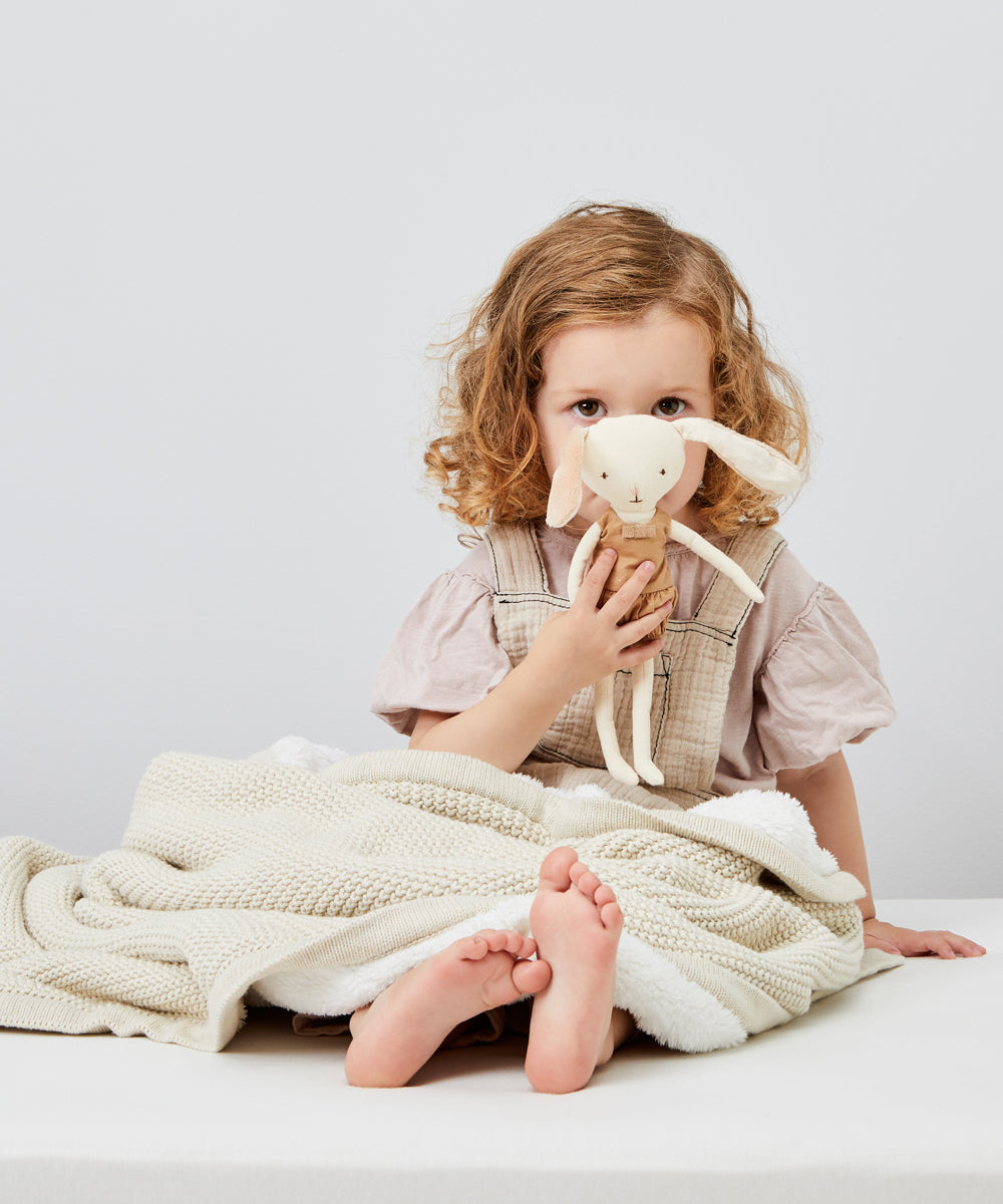 A child holding a soft toy sitting on teh floor with a linen coloured Little Green Sheep Organic Knitted Fleece Baby Blanket over heir knees