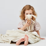 A child holding a soft toy sitting on teh floor with a linen coloured Little Green Sheep Organic Knitted Fleece Baby Blanket over heir knees