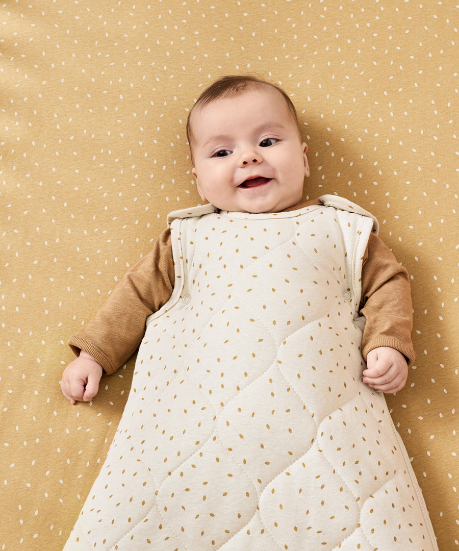 A baby wearing a linen rice print baby sleeping bag. The baby is lyinh down on a honey rice coloured cot sheet 