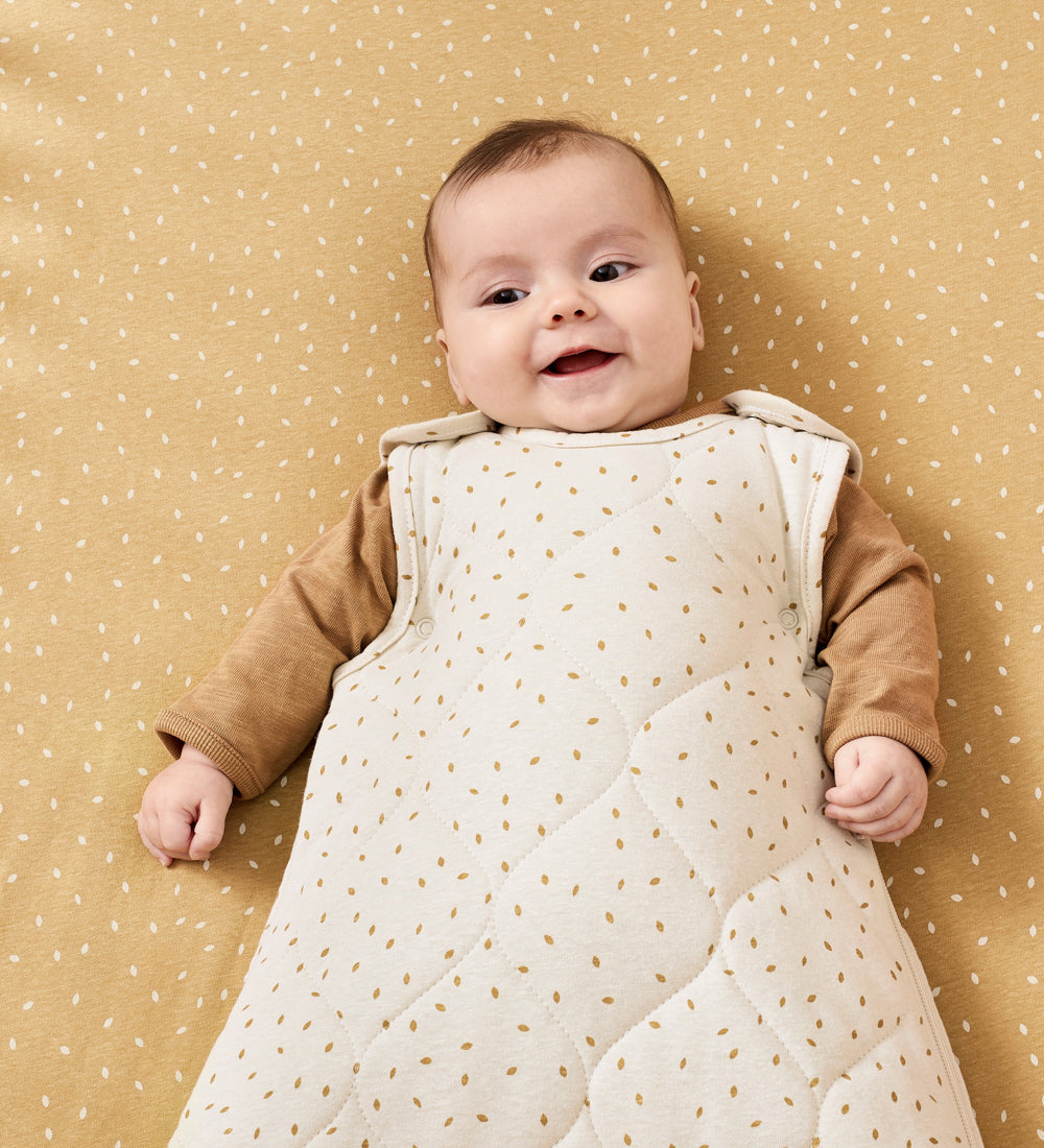 A baby wearing a linen rice print baby sleeping bag. The baby is lyinh down on a honey rice coloured cot sheet 