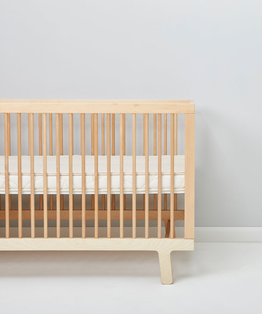 A cot pictured in a room in front of a light grey coloured wall. A Little Green Sheep Organic Cot Mattress Protector can be seen placed on the mattress inside the cot 