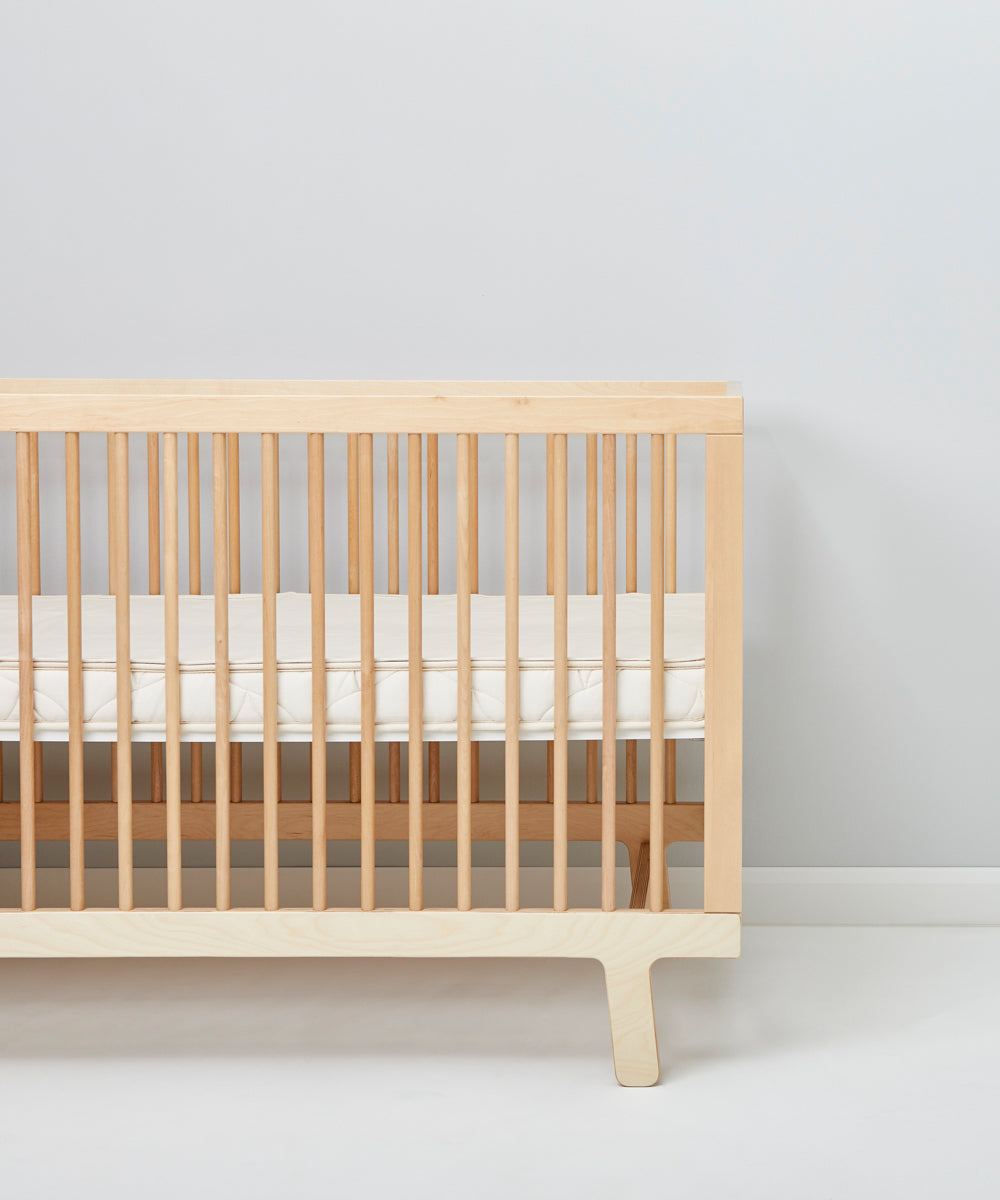 A cot pictured in a room in front of a light grey coloured wall. A Little Green Sheep Organic Cot Mattress Protector can be seen placed on the mattress inside the cot 