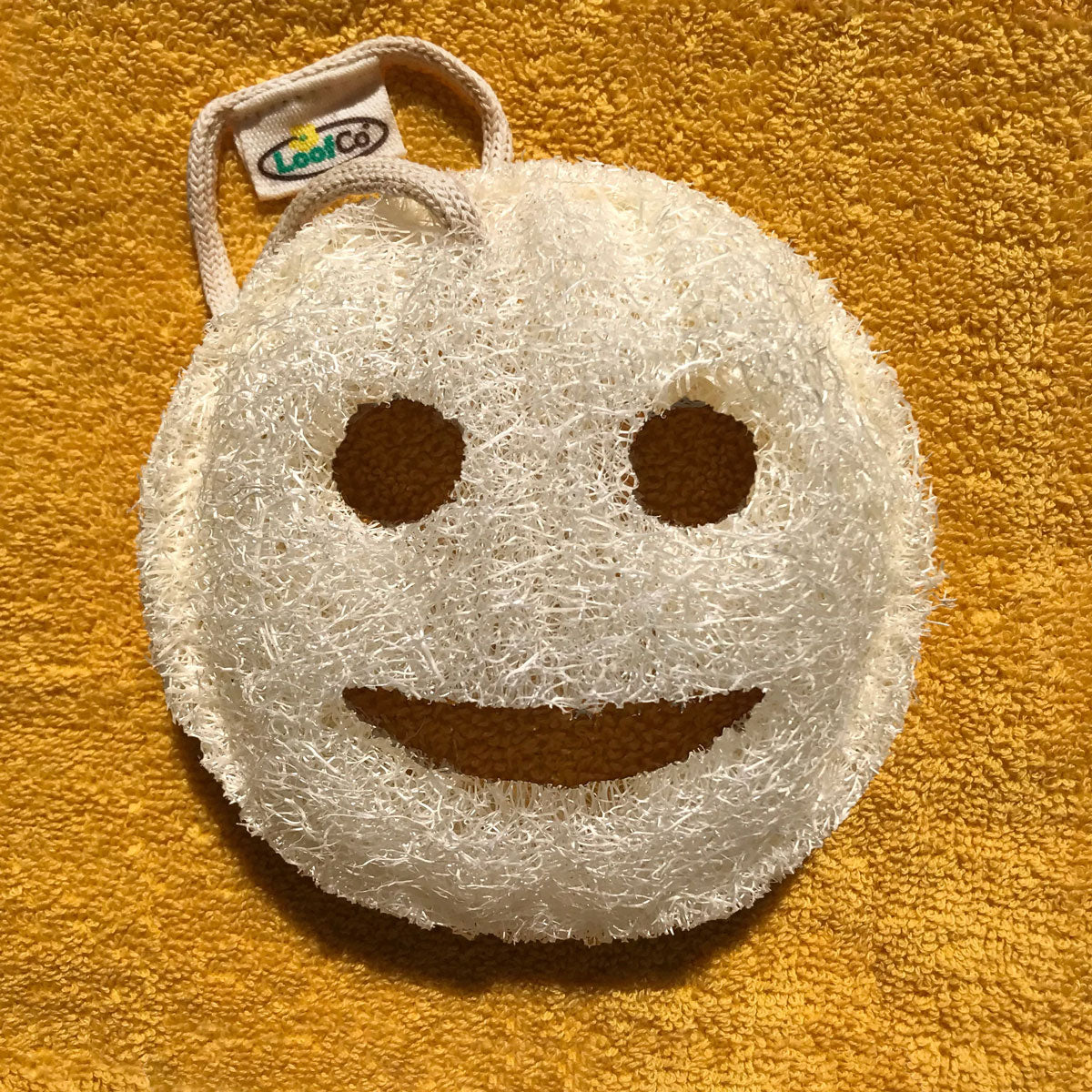 LoofCo Bath Time Smile Loofah pictured on a yellow coloured towel