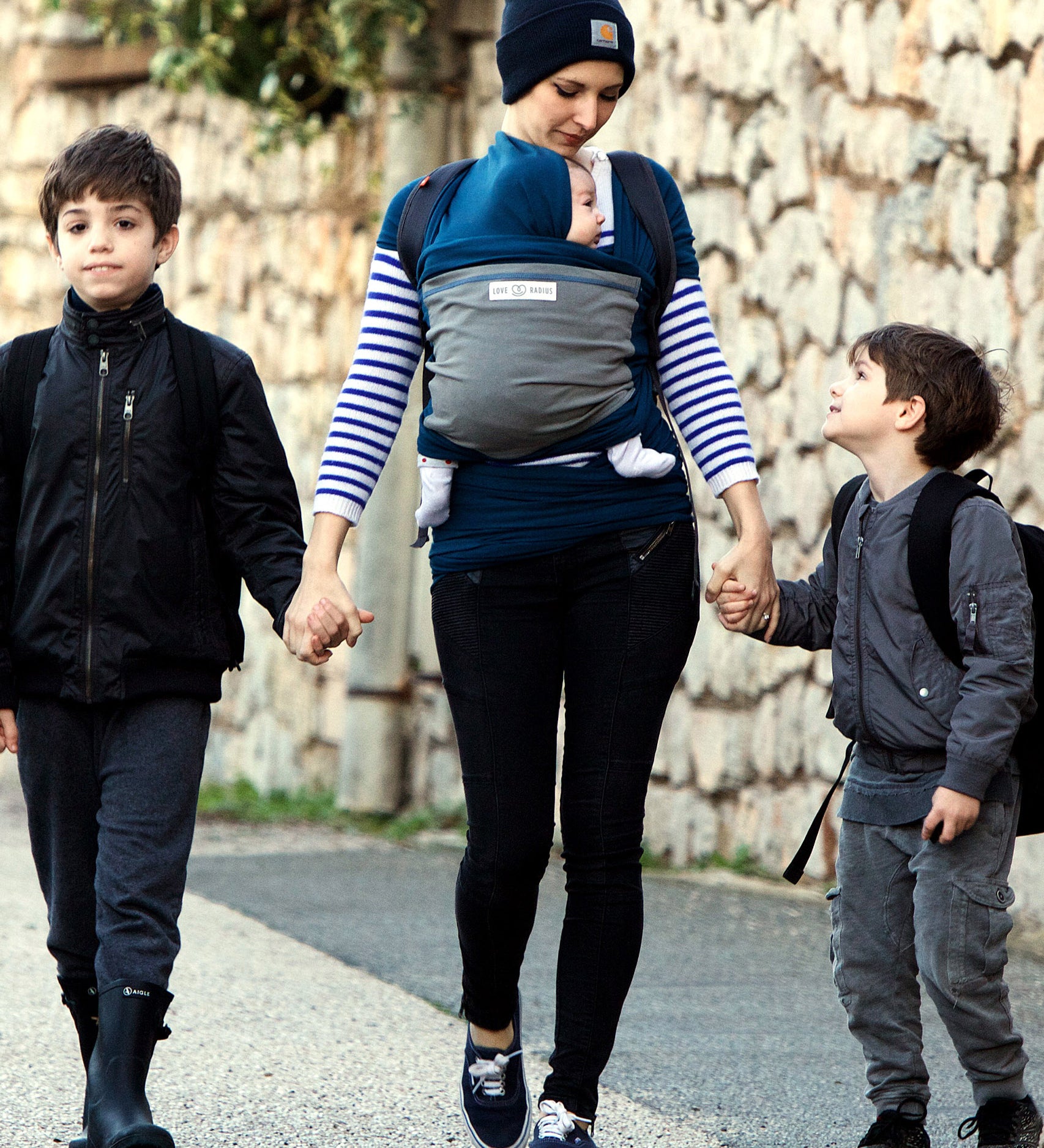 Adult carrying a baby in the Love Radius Original JPMBB Baby outside holding the hands of two younger siblings.