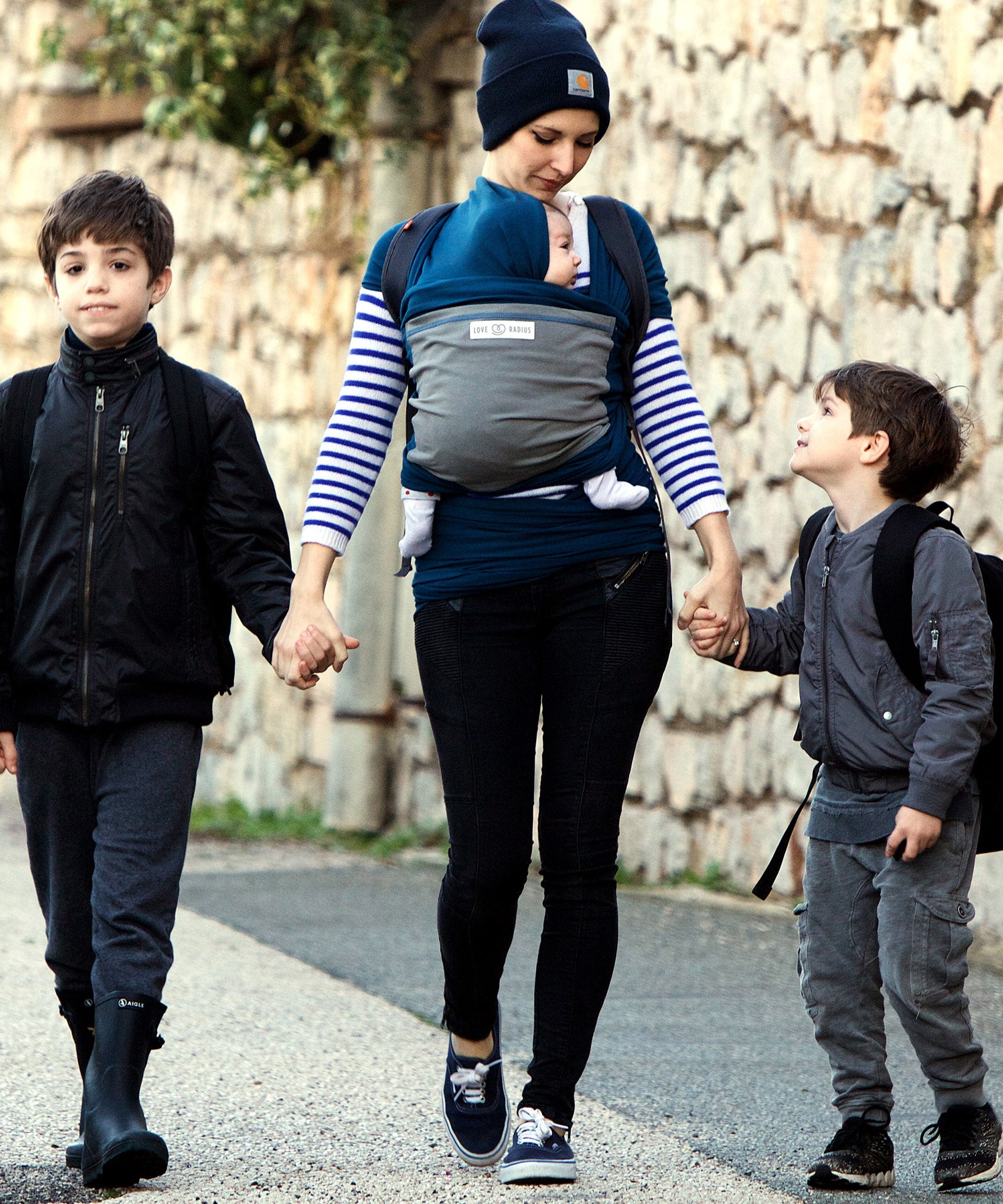 Adult carrying a baby in the Love Radius Original JPMBB Baby outside holding the hands of two younger siblings.