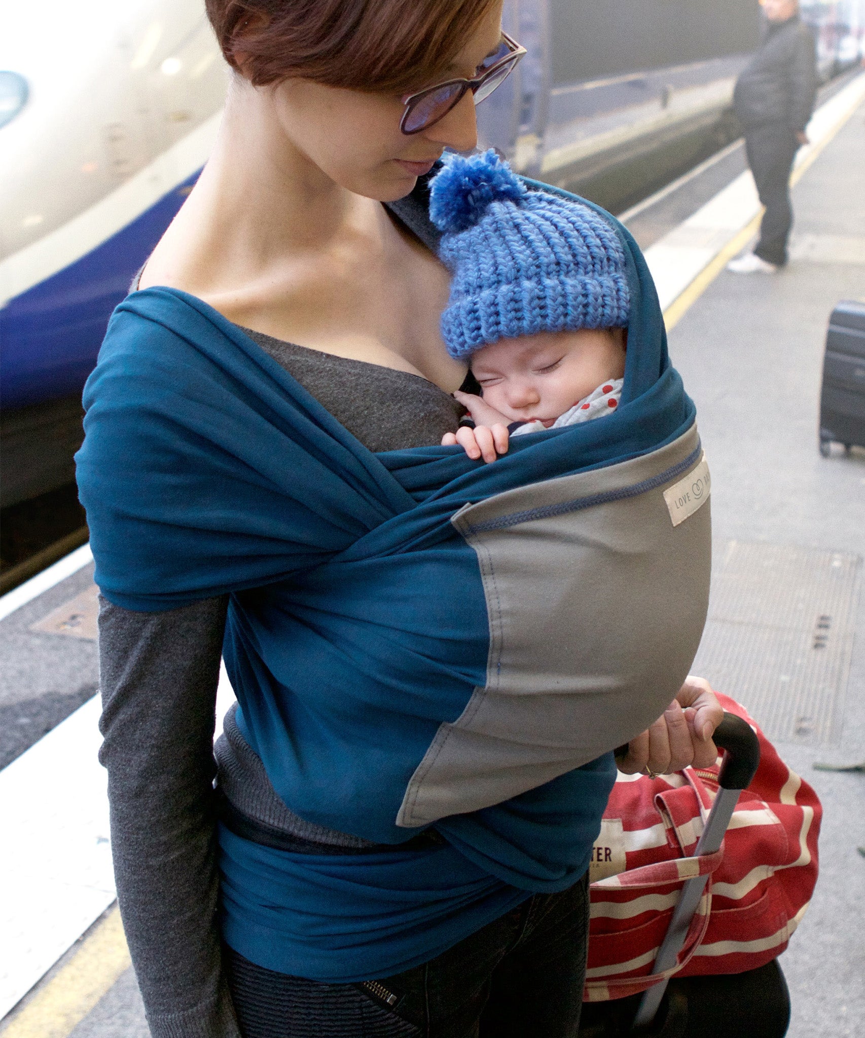 Adult carrying a baby in the Love Radius Original JPMBB Baby Wrap - peacock blue/elephant at a train station