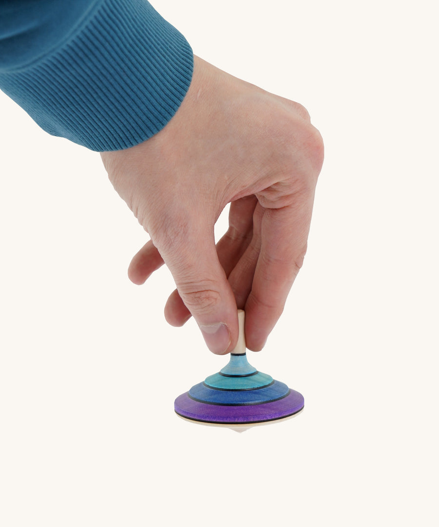 A wooden purple and blue flamenco Mader spinning top held by a hand on a cream background.