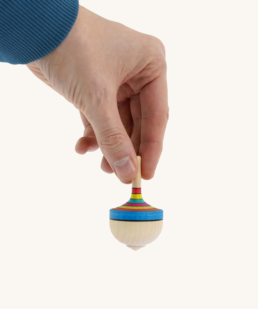 A wooden Mader traditional rainbow spinning top held by a hand on a cream background.