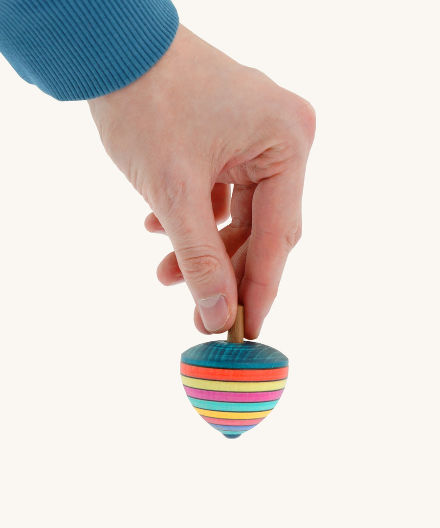 A wooden Mader dragon egg spinning top with multicoloured stripes  held by a hand on a cream background