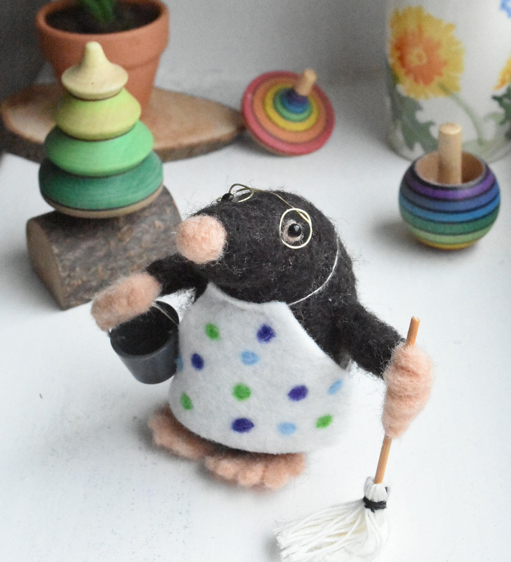 A felted mole figure wearing a spotty apron holding a bucket and a mop. The figure has been created using The Makerss Small Busy Mr Mole Needle Felt Kit.