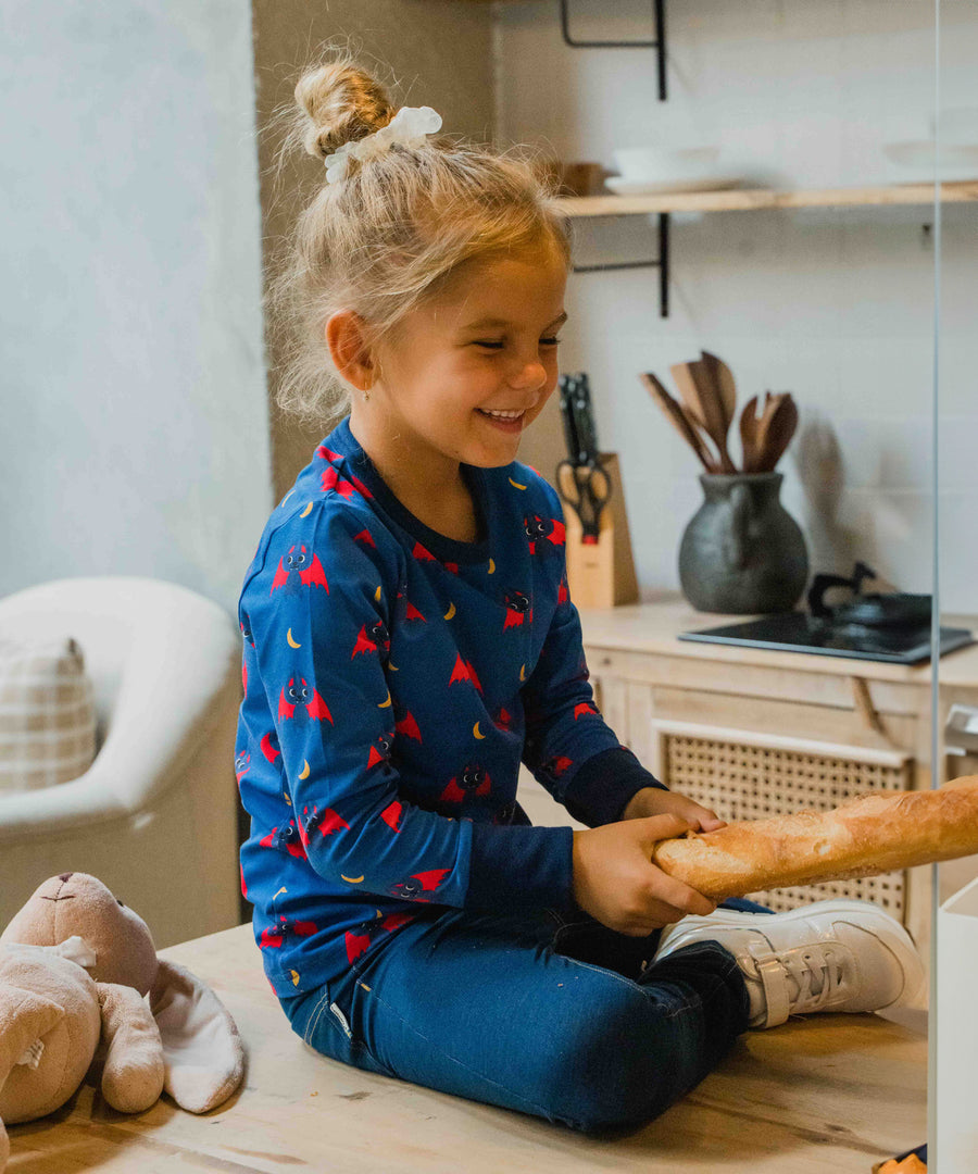 A child wearing the Maxomorra bat print top. The child is sitting on a wooden surface holding a baguette. 