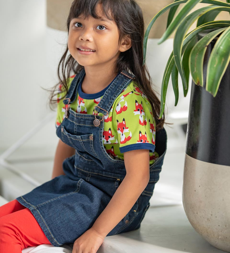 Young girl sat down wearing the Maxomorra eco-friendly navy denim pinafore dress over the short sleeve fox top