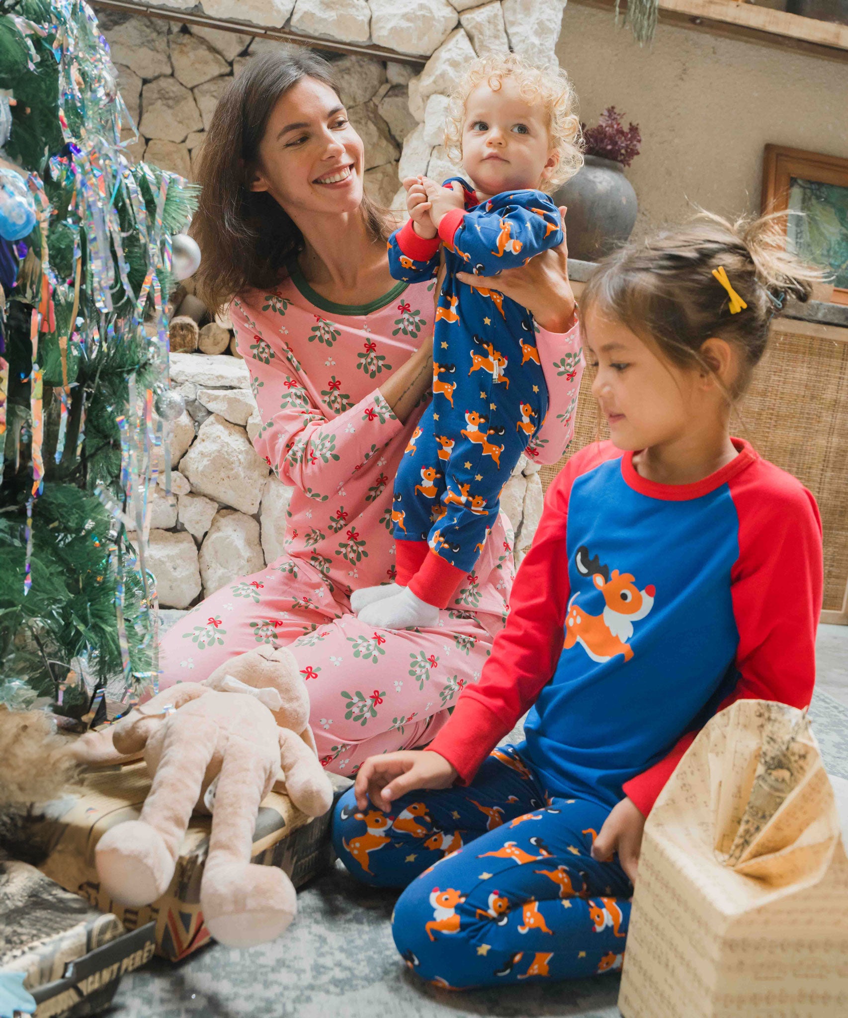 A young child wearing the Maxomorra Reindeer Long Sleeve Rompersuit being held by a woman wearing pink pyjamas. Another child sits in front of them in reindeer print leggings and a matching reindeer raglan top. 