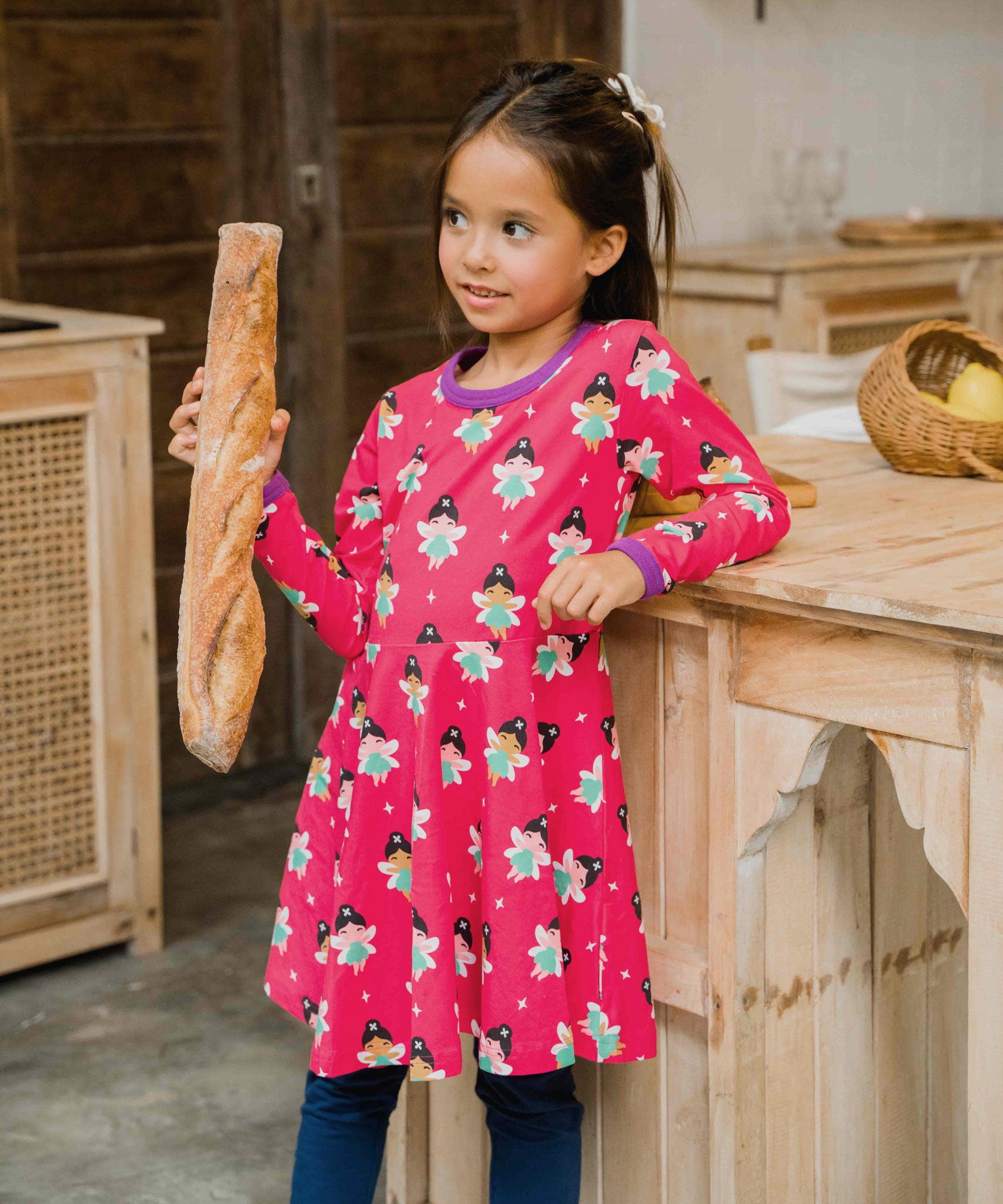 A child wearing the Maxomorra Fairy print circle dress. The child is holding a baguette and is leaning against a kitchen counter top 