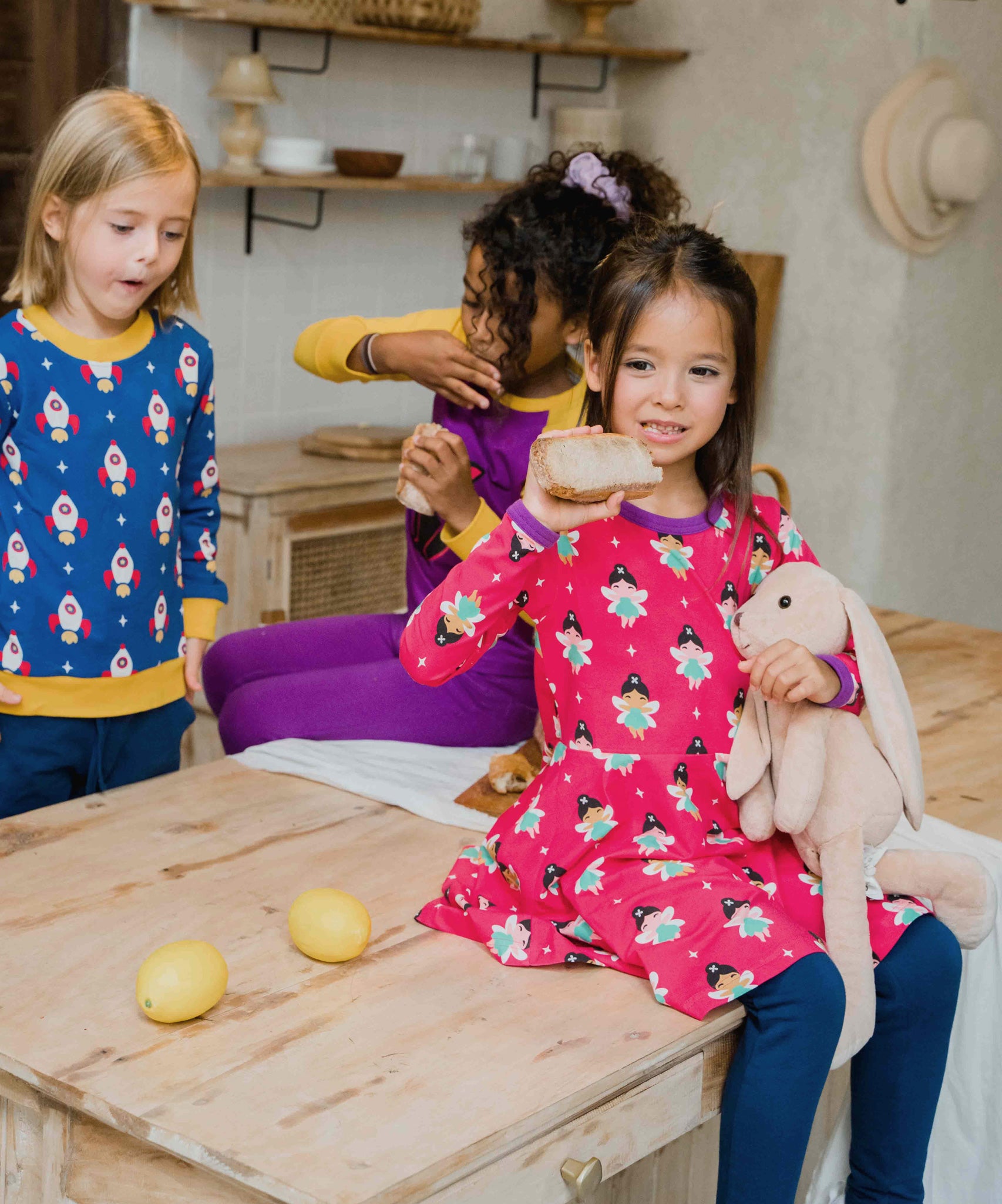 A child wearing the Maxomorra Fairy print circle dress. The child is sitting on a kitchen counter top. Other children can be seen in the background. 